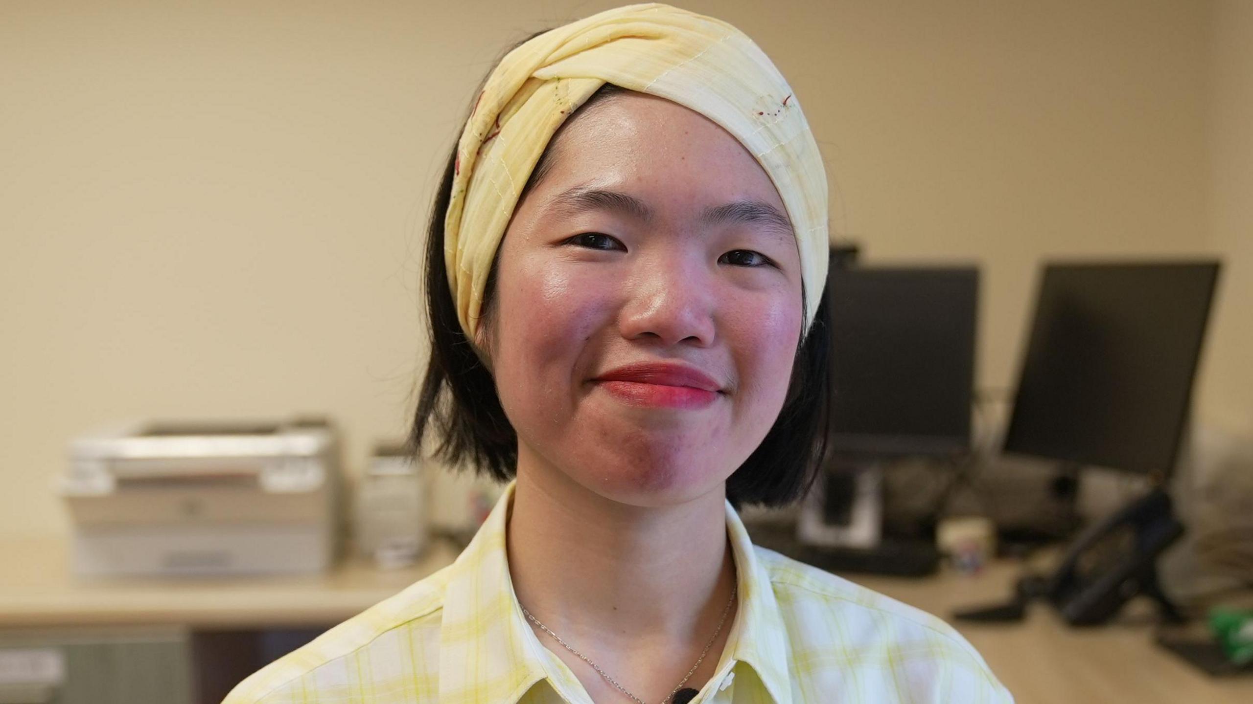 Dr Gillian Chin is wearing a checked yellow shirt and headband.  She is in her GP surgery  - Erskine Medical Practice in Dundee