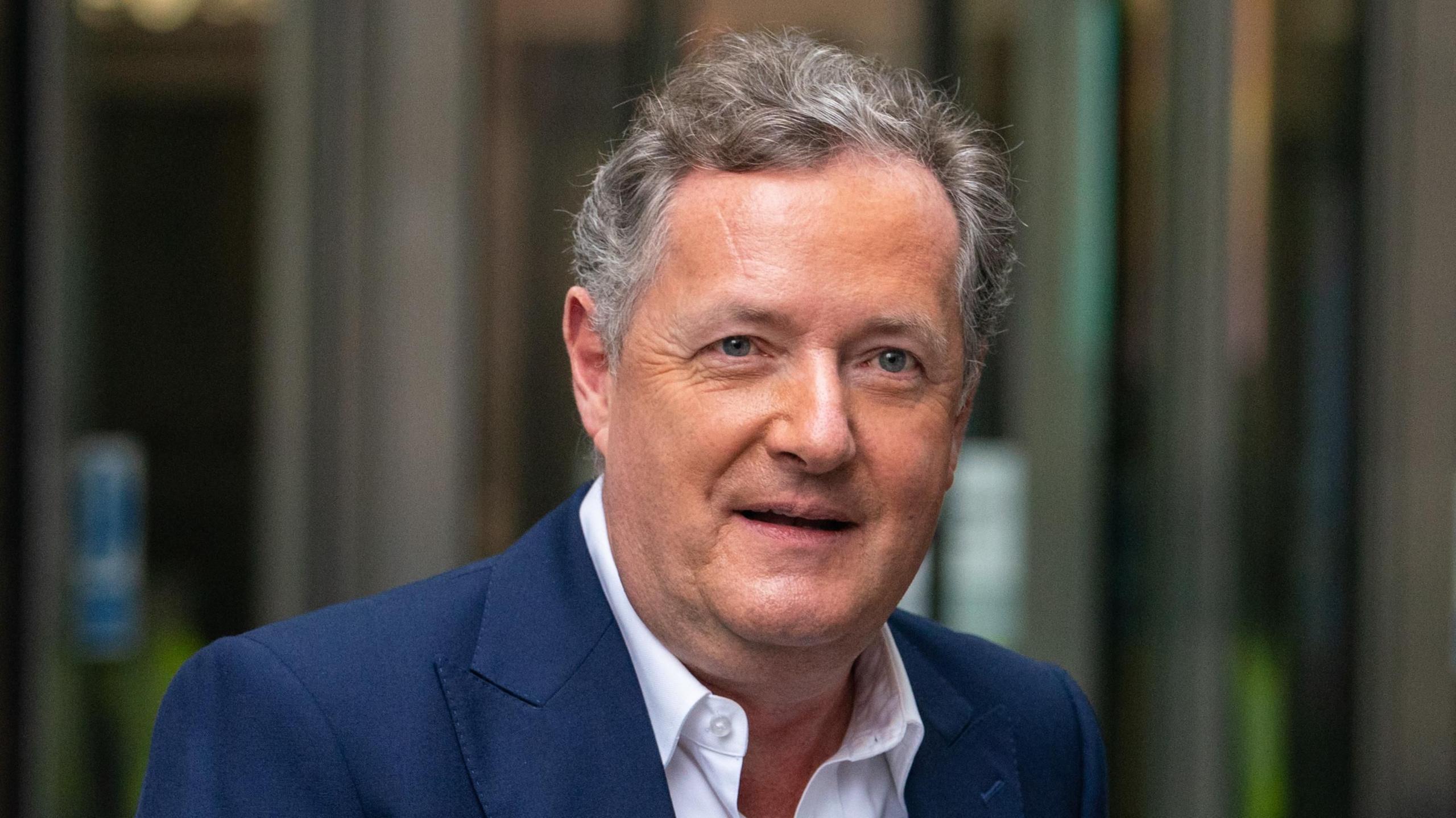 Piers Morgan, a man with short grey hair and wearing a blue suit with a white shirt undone at the collar, looks ahead