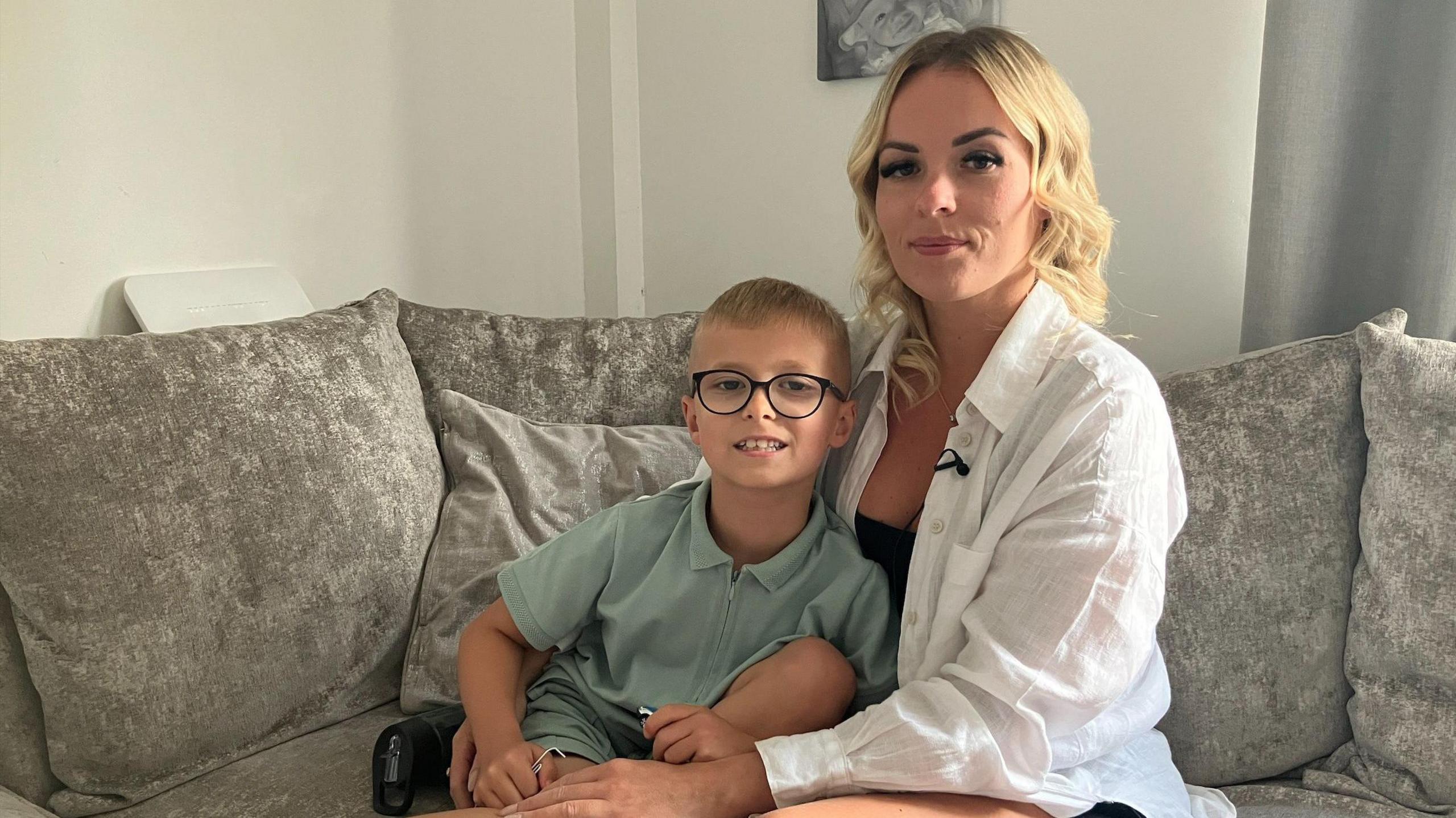 Alison McKay and her son Freddie sit on a grey sofa together. Both are looking at the camera and smiling slightly