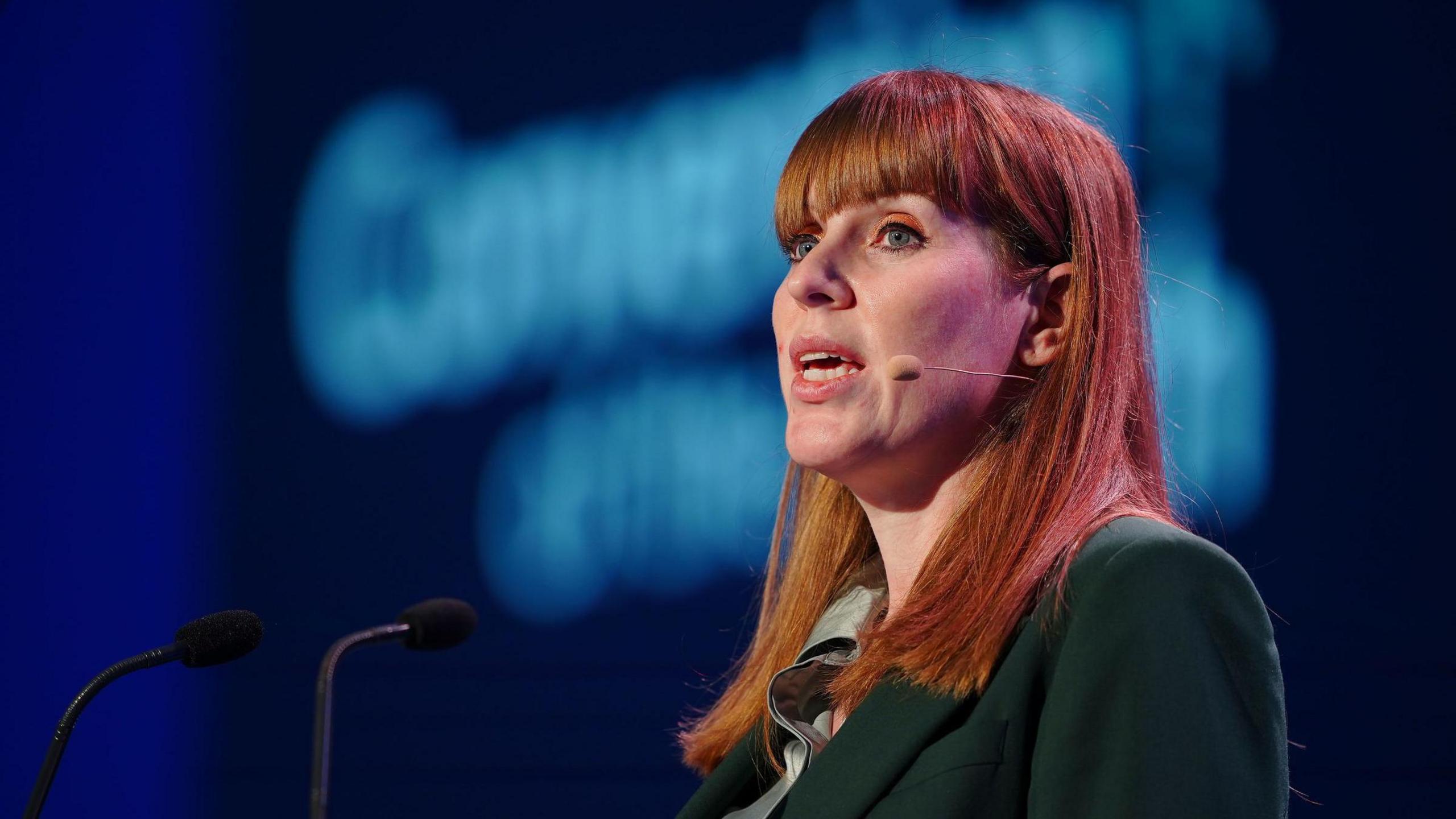 Angela Rayner is standing on a stage with two microphones in front of her. She's also wearing a microphone, along with a green jacket.