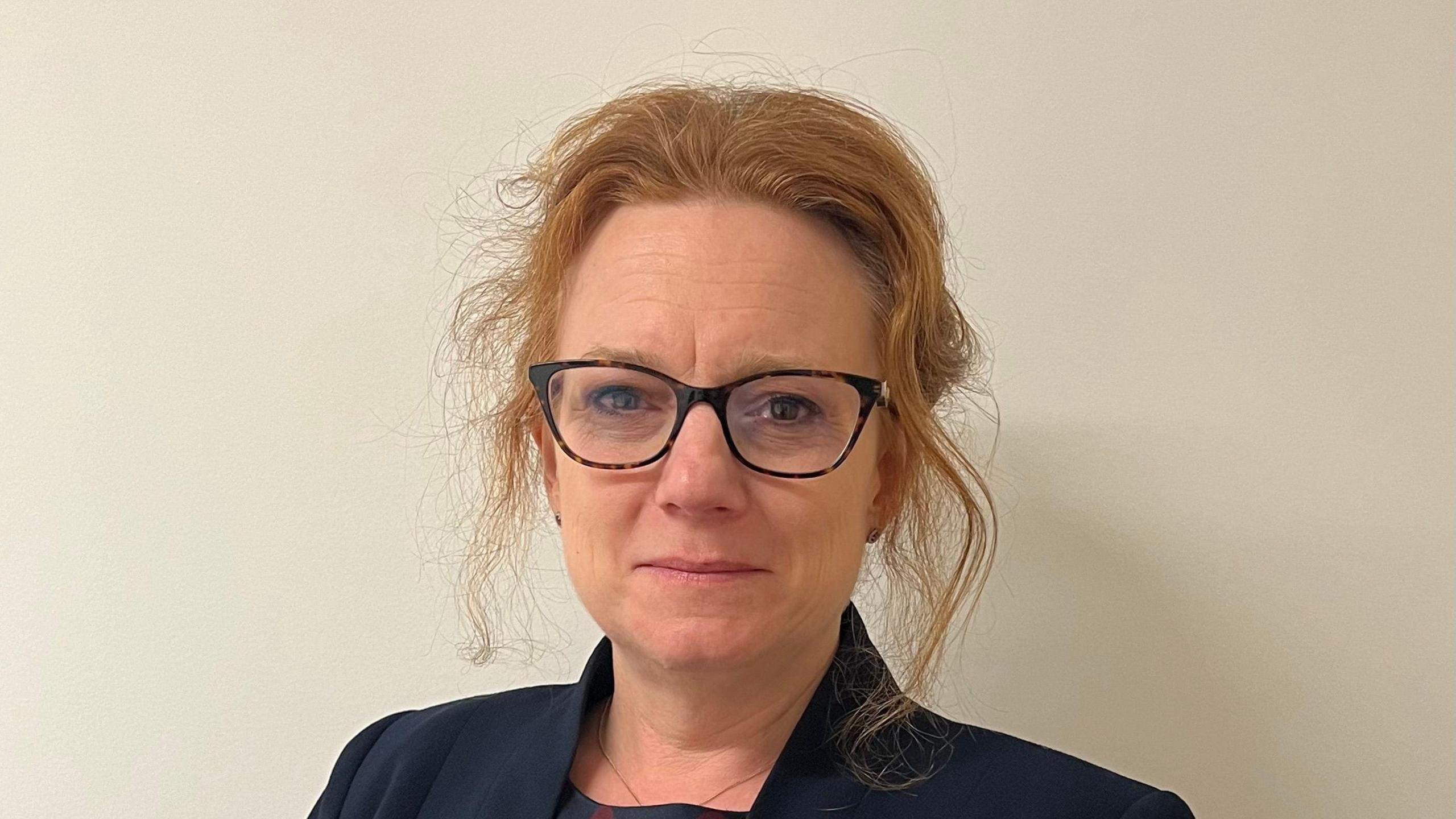 Sarah-Jane Smedmor smiles at the camera. She has red hair that has been tied back with some loose bits framing her face. She wears glasses and a navy jacket.