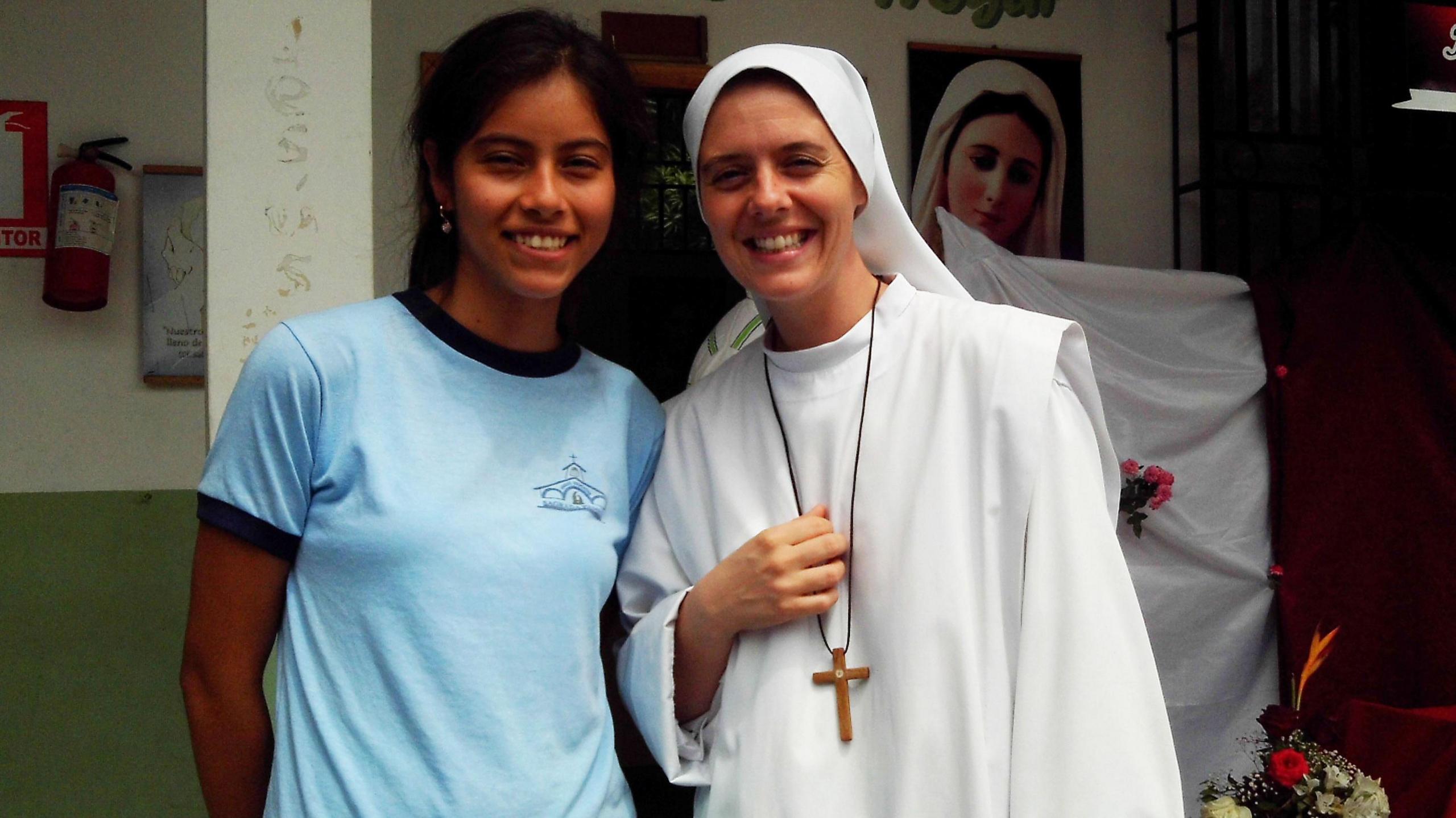 Sr Clare in a white habit and wearing a long black necklace with a wooden cross stands beside a young girl in front of a white and green wall.