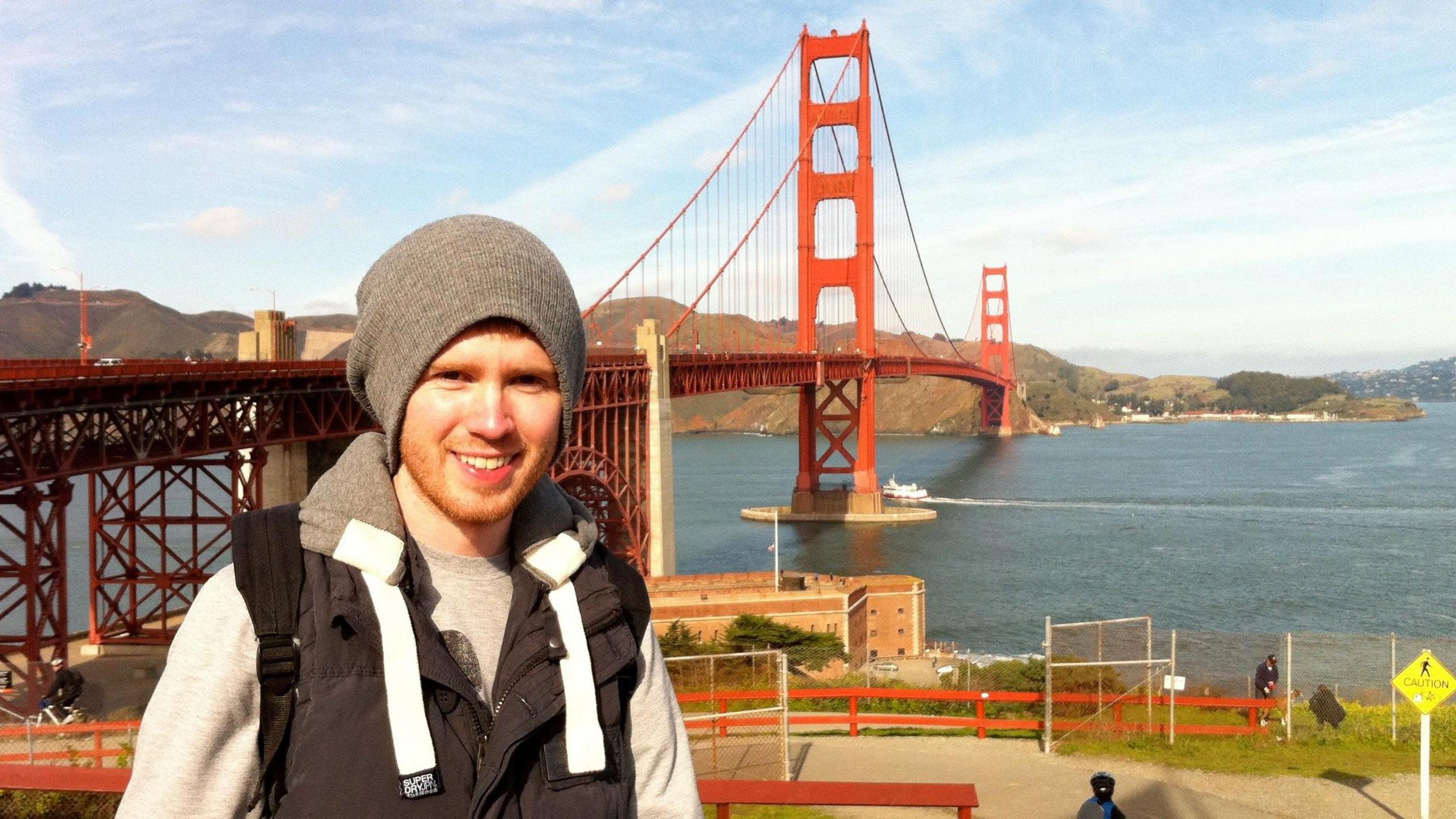 A young Jack is in San Francisco. The famous Golden Gate Bridge is behind him. He wears a grey hoodie, black sleeveless jacket, and grey beanie. He has a ginger beard.