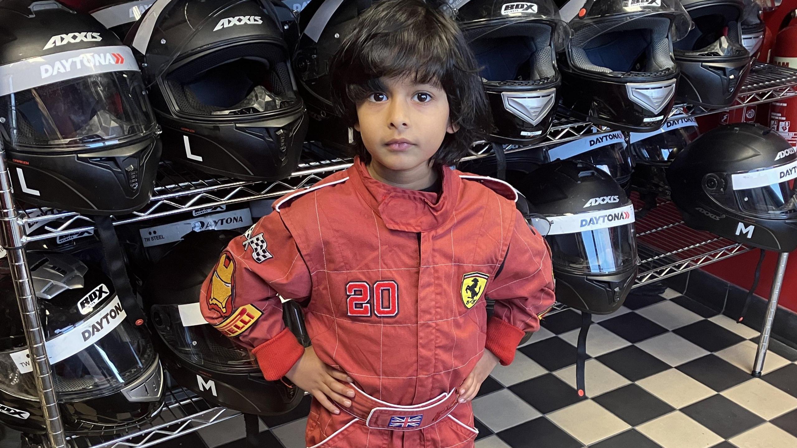 Raef is standing on a black and white chequered floor, with a large stand behind him that holds black racing helmets. He has black hair and is wearing a red racing suit with the Ferrari F1 logo on it.