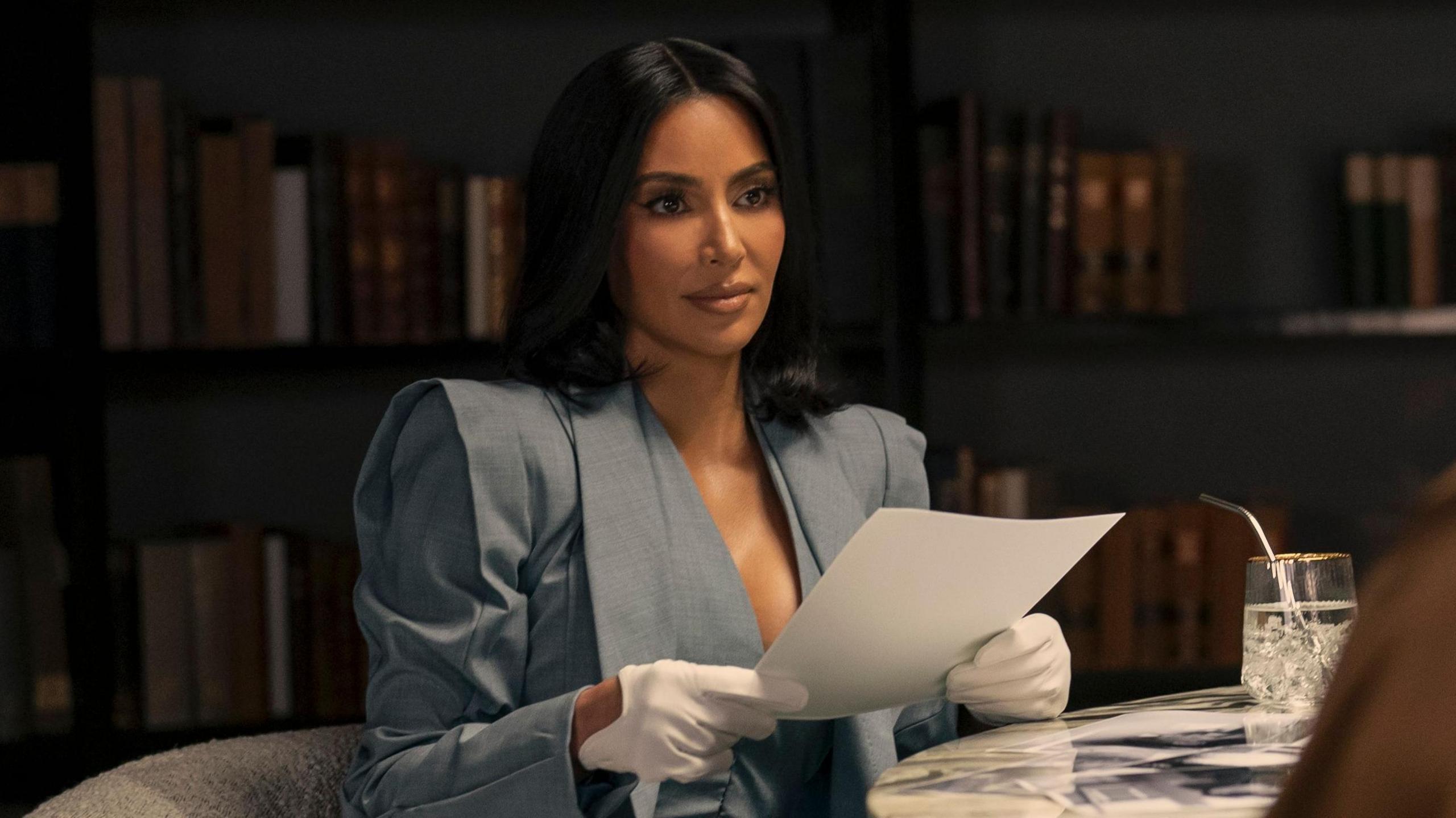 Picture of Kim Kardashian starring in All's Fair on Disney. She is wearing a grey suit jacket with huge shoulder pads and white gloves.