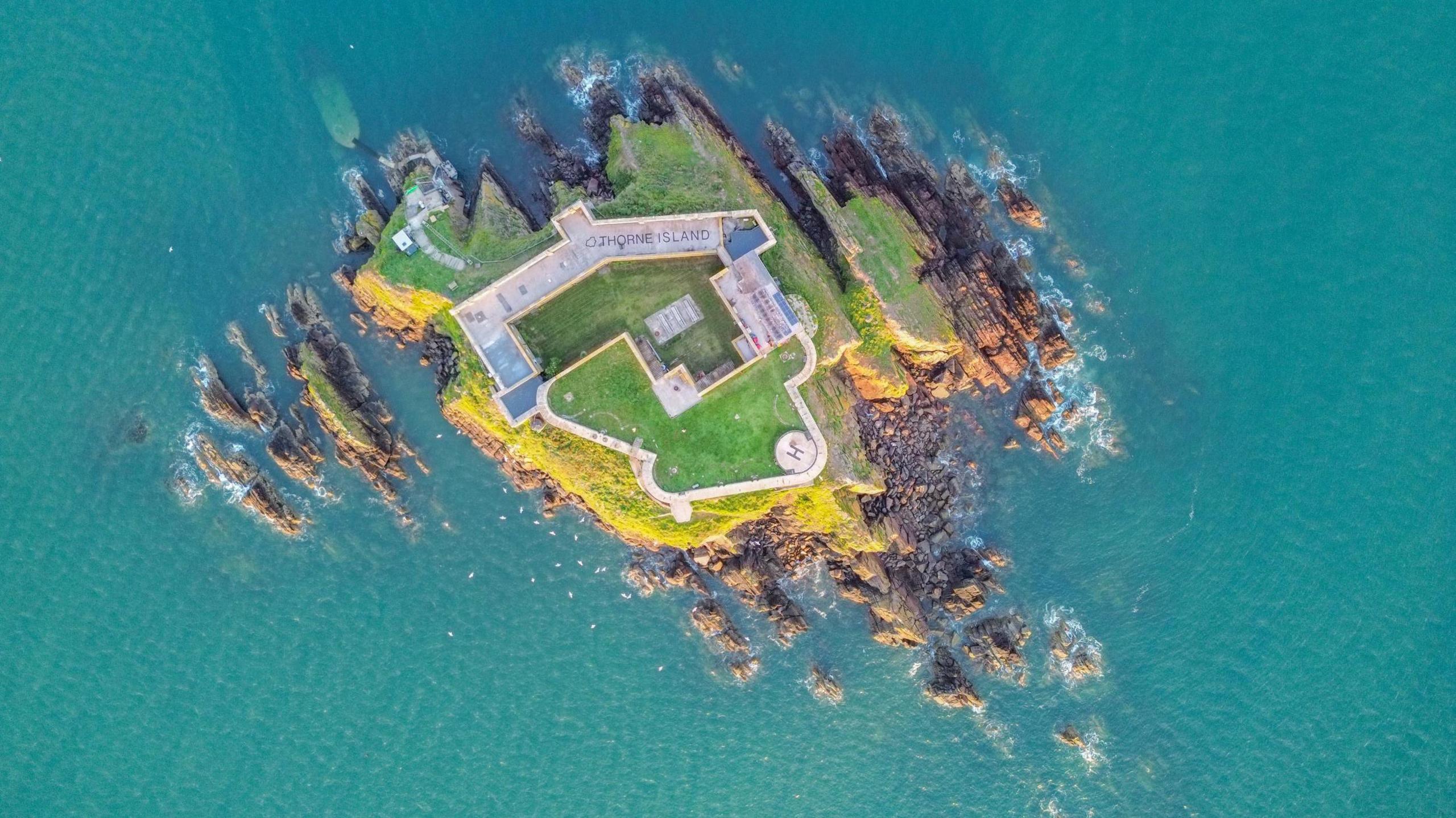 An aerial view looking directly down on to the island. The stone outer walls surround the majority of the land, with a green courtyard inside. Cliffs and rocks surround it, the sea is turquoise blue. 