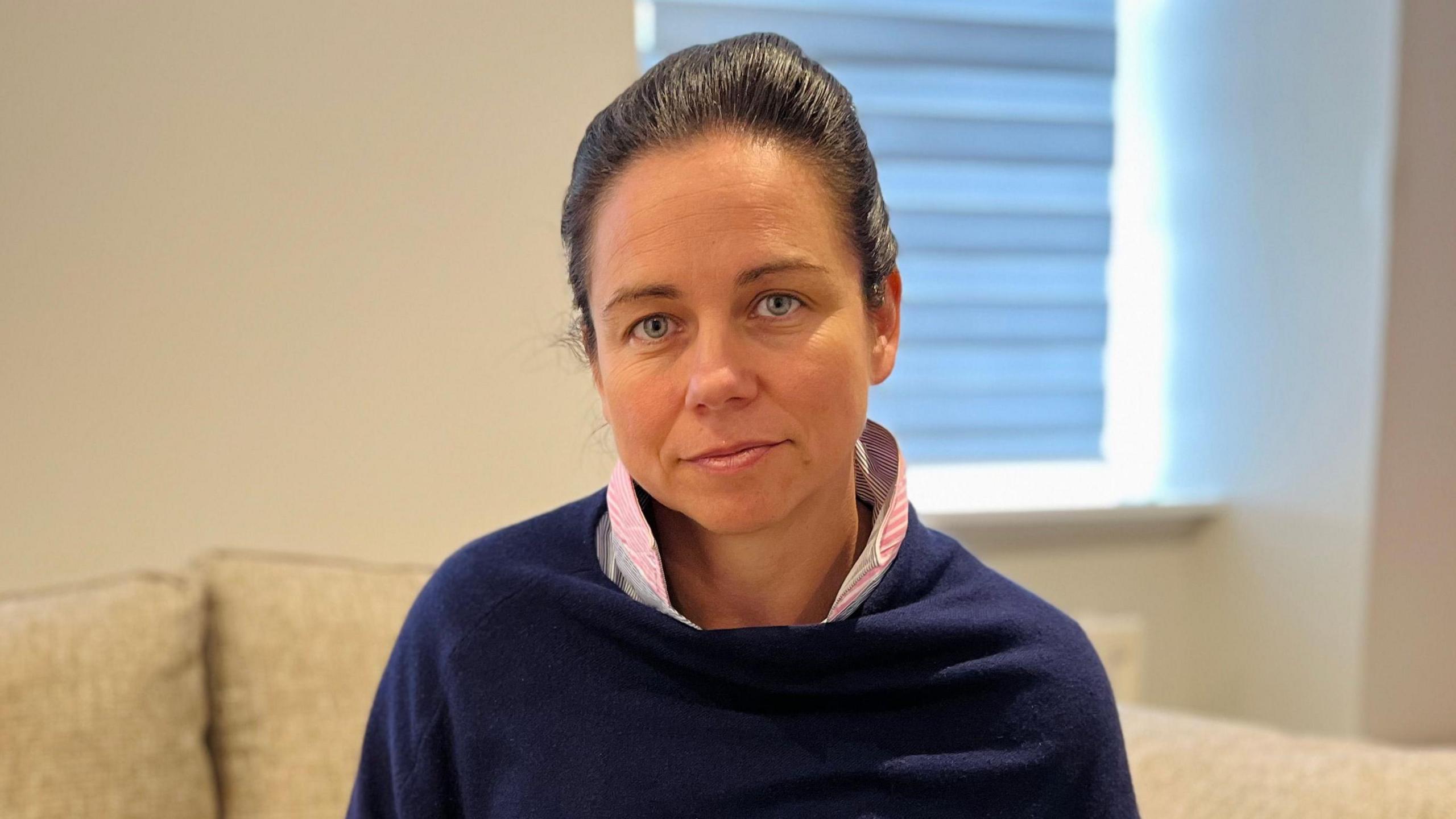 A woman in a blue jumper with a pink shirt underneath looks into the camera while seated on a sofa