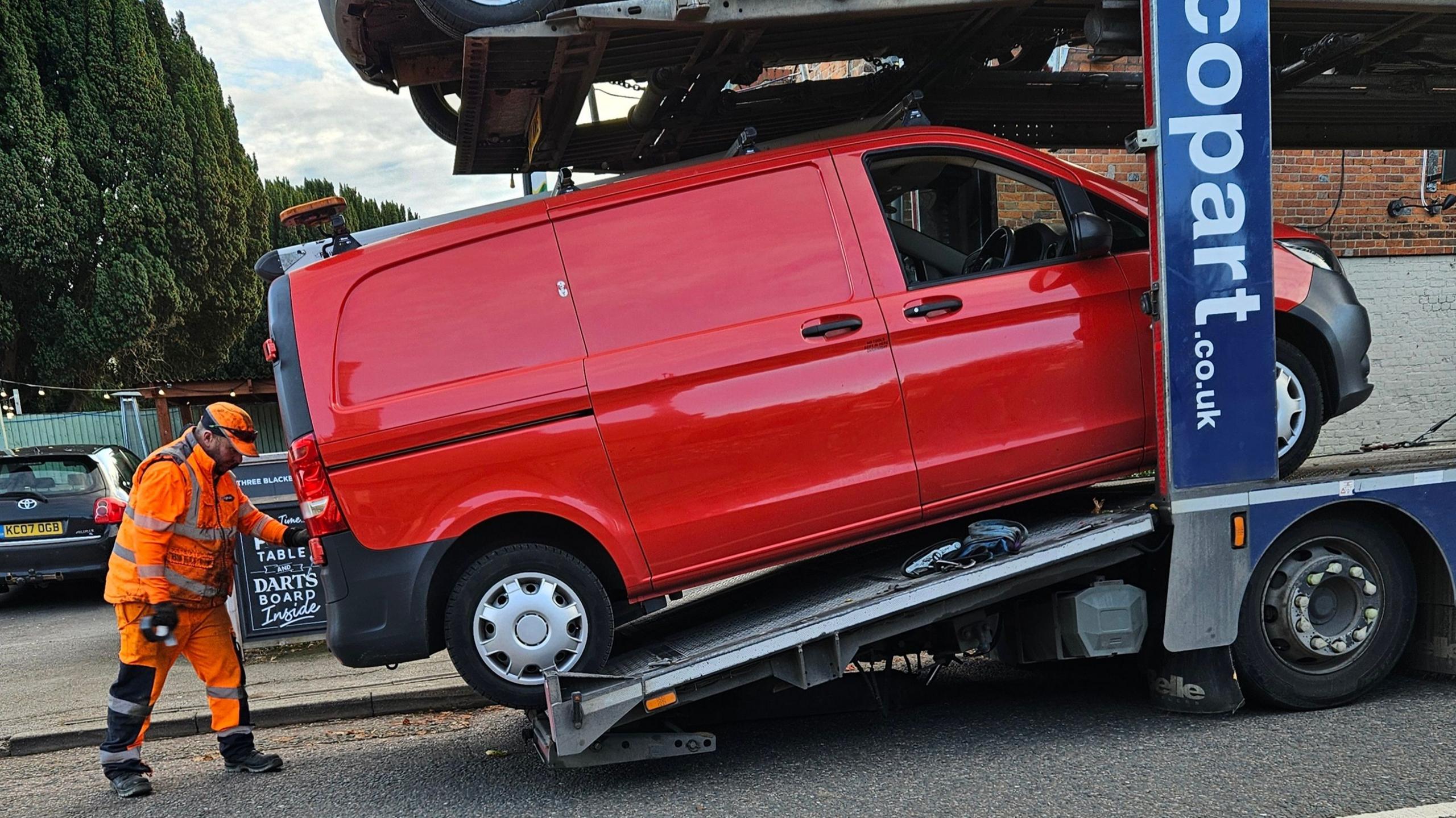 A red van being loaded on to a low loader by a man in a fluorescent orange outfit