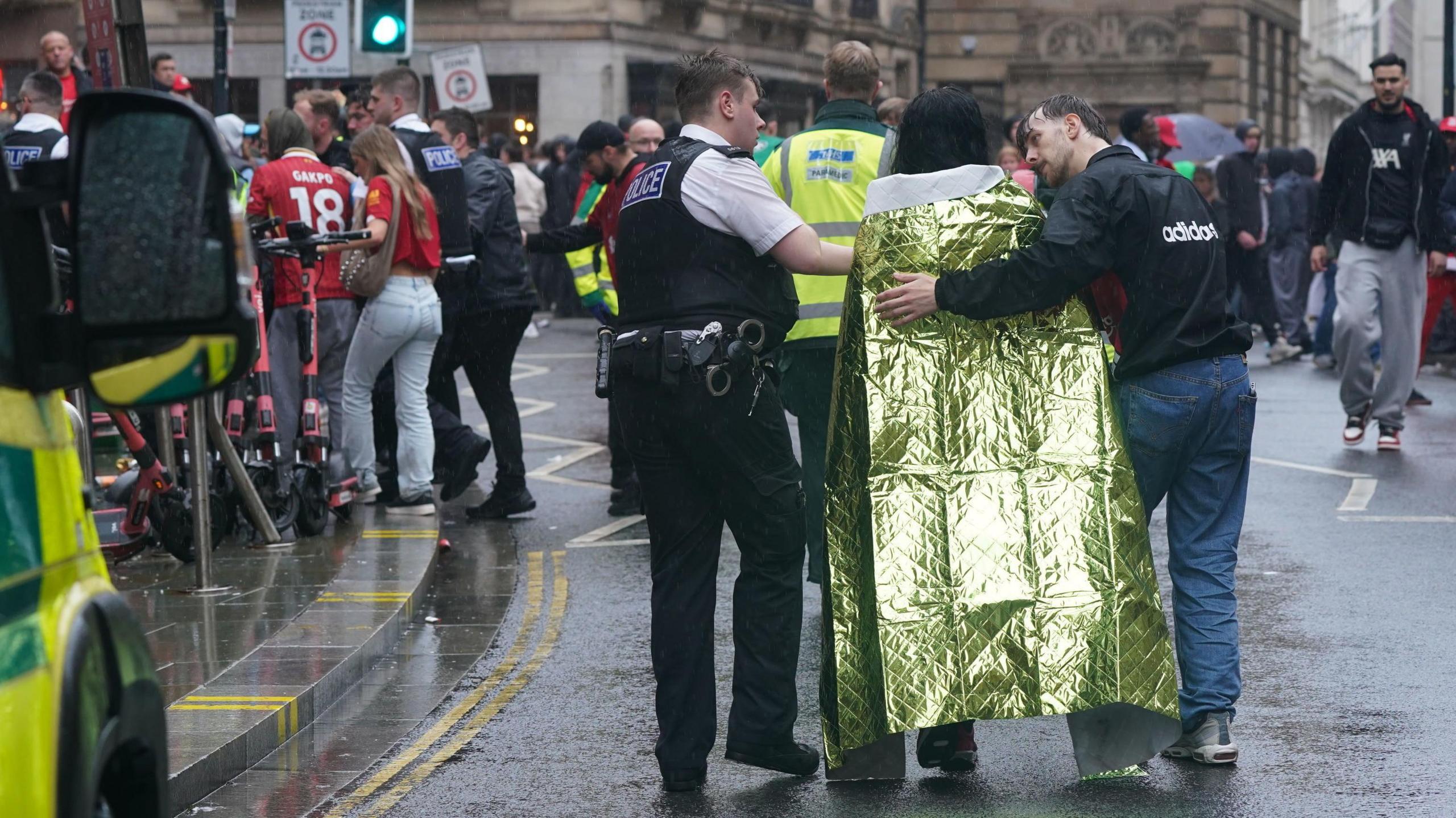 A police officer helps a woman wrapped in foil blanket walk down the road, and a man in a black jacket has his arm wrapped around their should as he talks to the woman