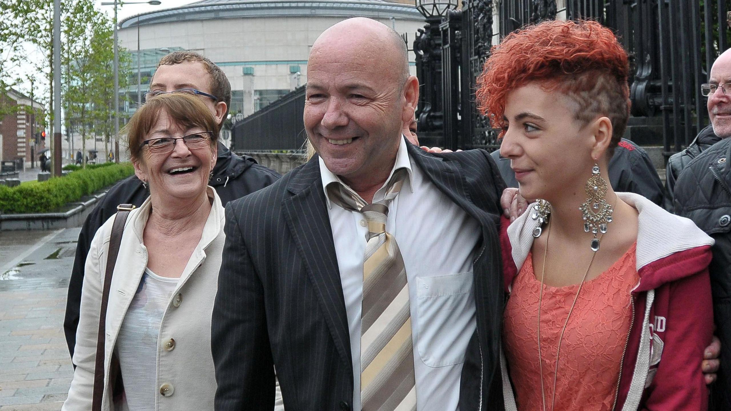 Belfast Court is behind with street trees, black gates and Liam holding centre, smiling with his family. 
He is bald, wearing a white shirt, black striped suit and beige stripy tie. 
The woman on his left is wearing a beige coat, white top and she has brown hair tied up with a fringe and brown handbag. 
The boy behind her's face isn't visible.
The young woman on the right of Holden has red crimped hair, tied up on one side and shaved into patterns on the other. She is wearing a pink hoodie and coral top with long dangly silver earrings and a long necklace.