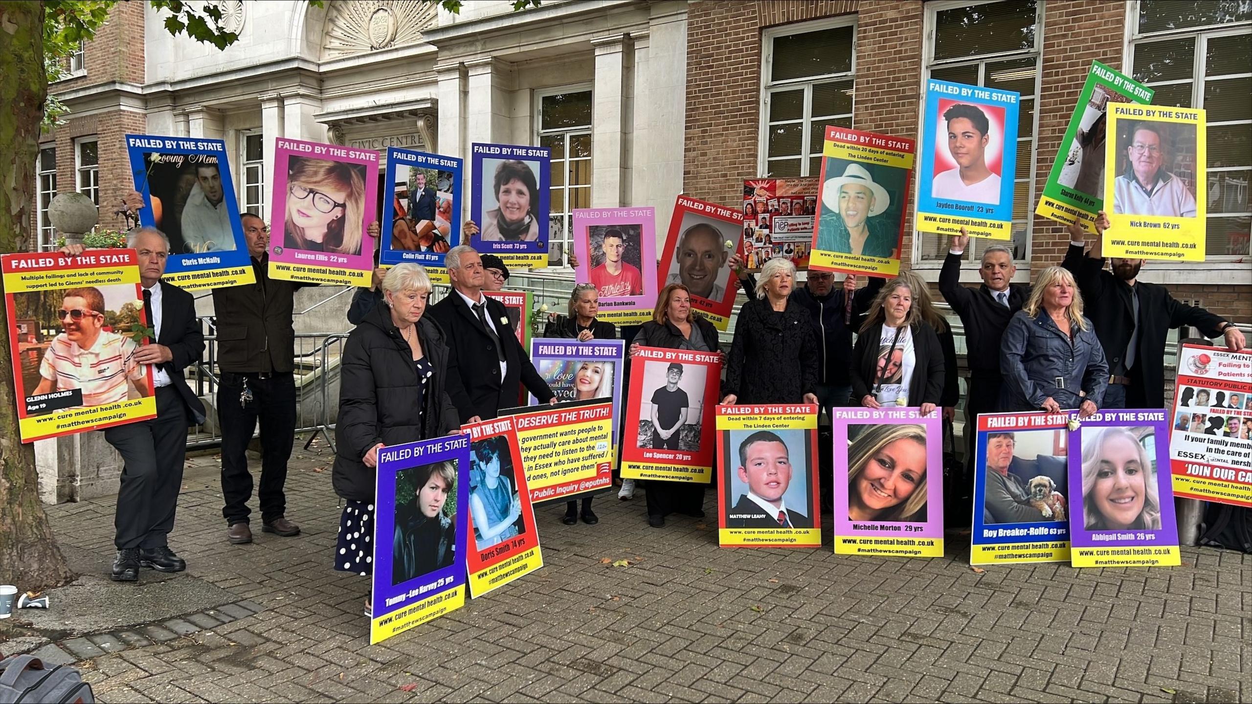 A large group of bereaved family members stand outside the Civic Centre in Chelmsford at the first day of The Lampard Inquiry holding large posters of their loved ones with captions such as "failed by the state". They are wearing coats and black clothing.