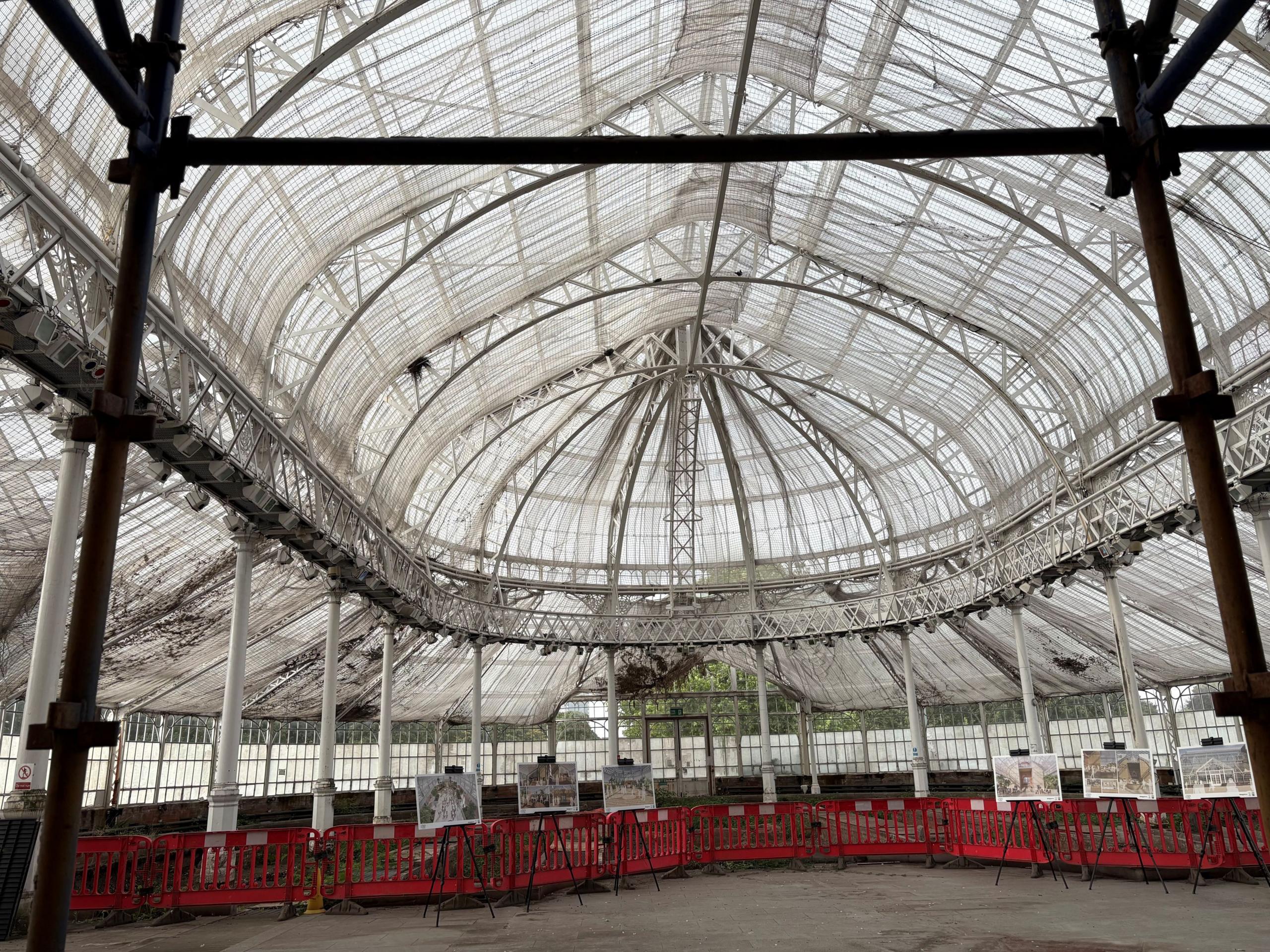 An image of a huge class house in daylight, clearly in the midst of construction work - with red barriers separating the floor area, and netting stretched across the ceiling under the glass panes.
