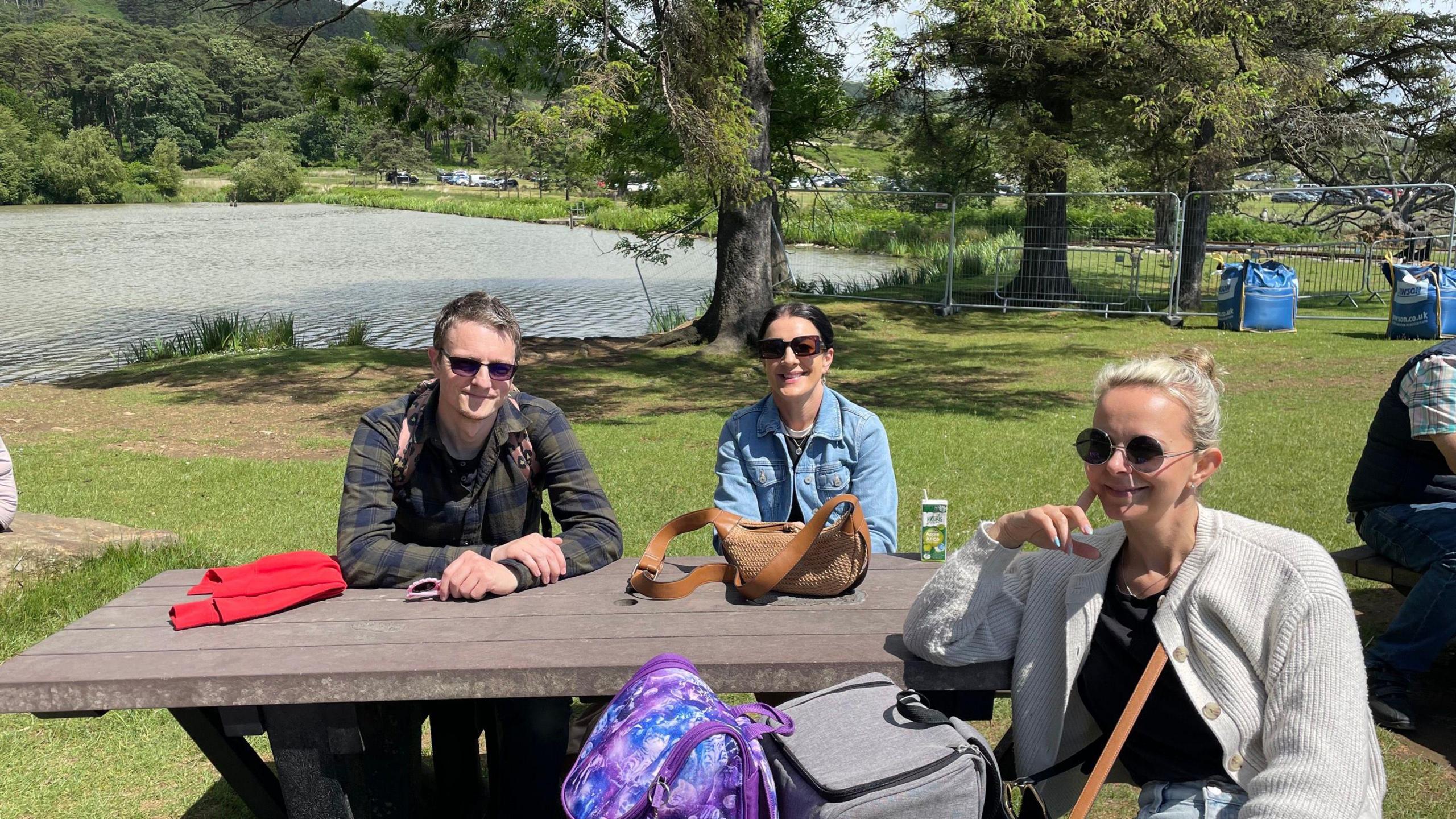 A man and two women sit on a brown wooden bench in front of a lake with trees behind them. They all wear sunglasses and smile at the camera. The man is wearing a blue and green checkered top, the woman next to him has a denim jacket on, and the woman in front of them has a white cardigan on with a black top underneath. 