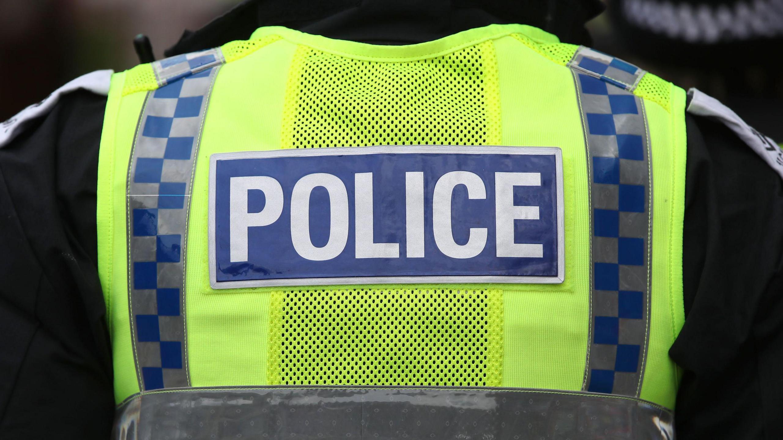 The back of a police officer's torso. They have a yellow high-vis on with the word police written in white on a blue rectangle background in the middle of the jacket.