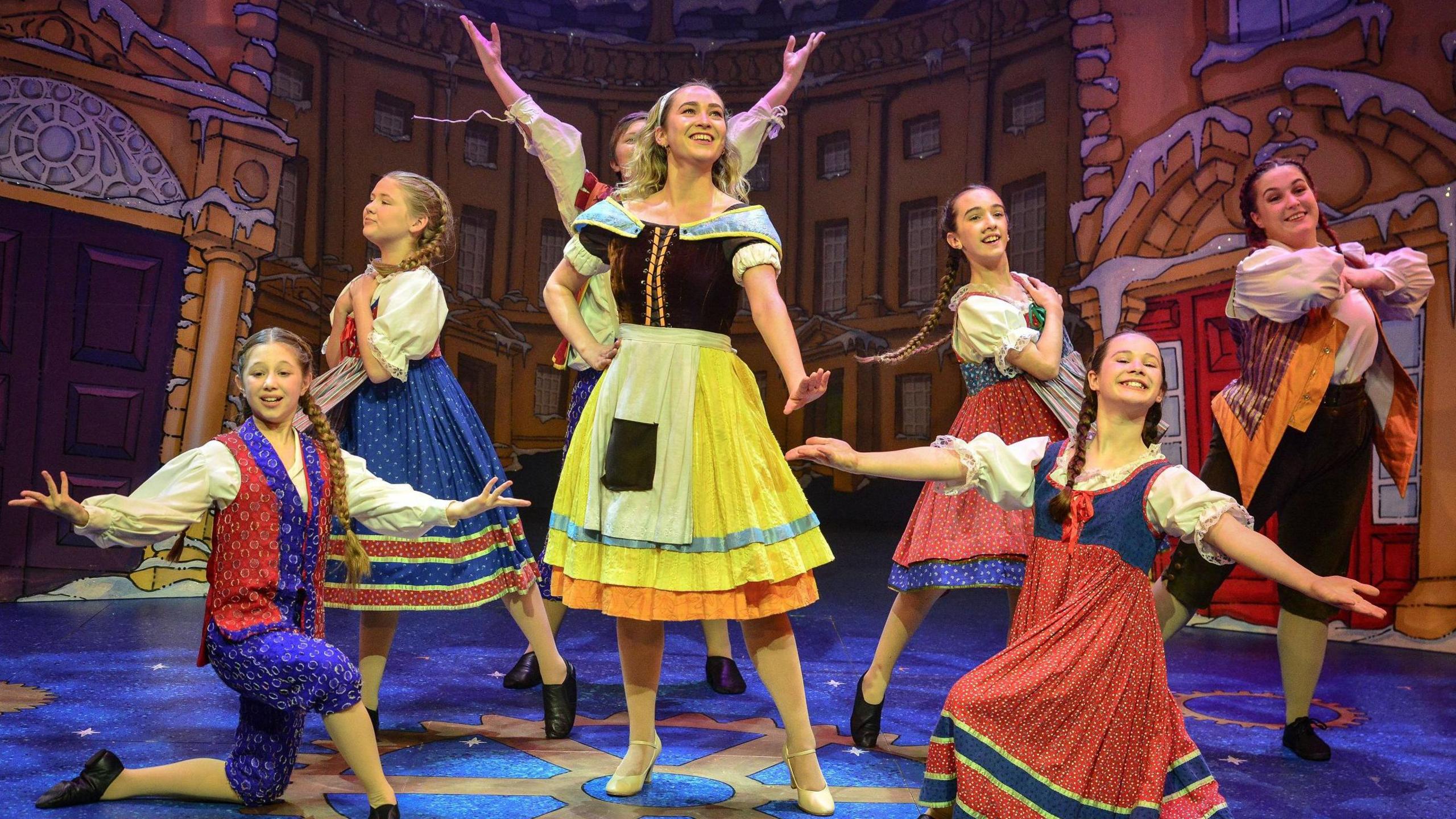 Seven performers on a stage, during a pantomime. The woman in the middle is dressed in a yellow skirt, with brown top, with five girl dancers around her with their arms out. There is a painted stage behind them. 