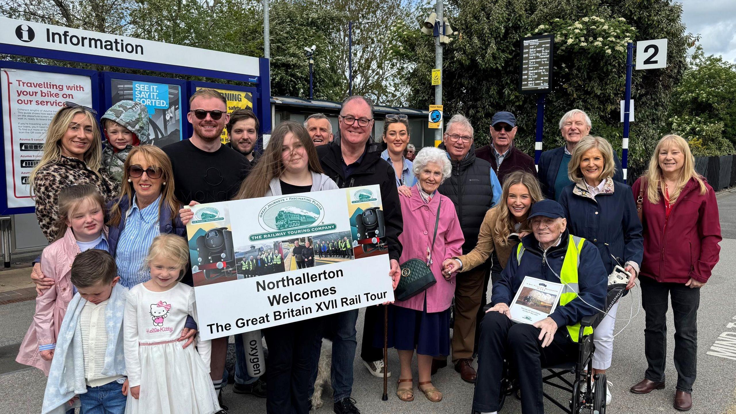 An elderly man in a wheelchair with his family. About 20 people including children. They are stood on a railway platform holding a sign that reads 'Northallerton Welcomes The Great Britain XVII Rail Tour'.