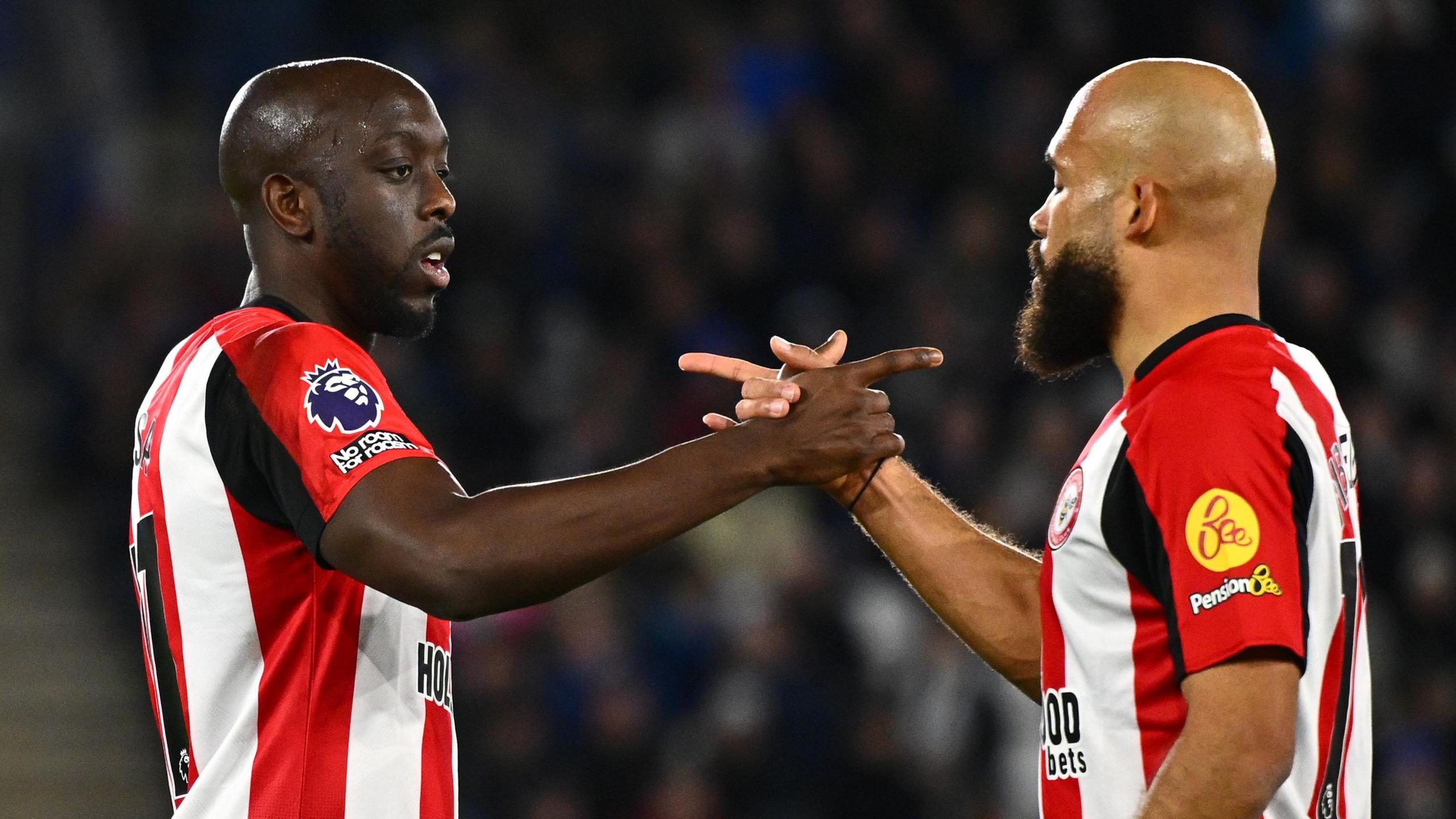 Yoane Wissa and Bryan Mbeumo, both wearing red and white striped Brentford home shirts, face each other and clasp hands while also using the index fingers of the same hands to point at each other during a game being played under floodlights