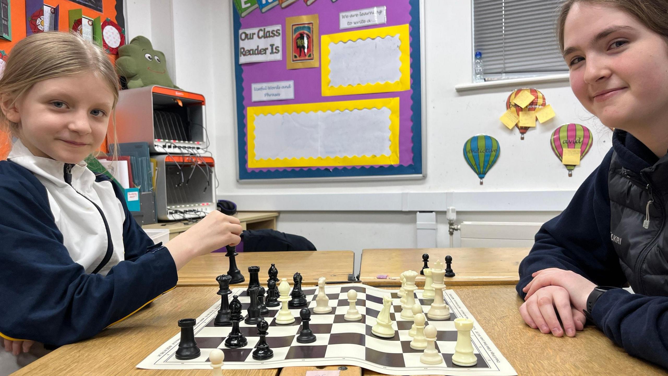 Two school girls, who are smiling at the camera, sit opposite one another. A chess board is in between then. They are sitting in a classroom.