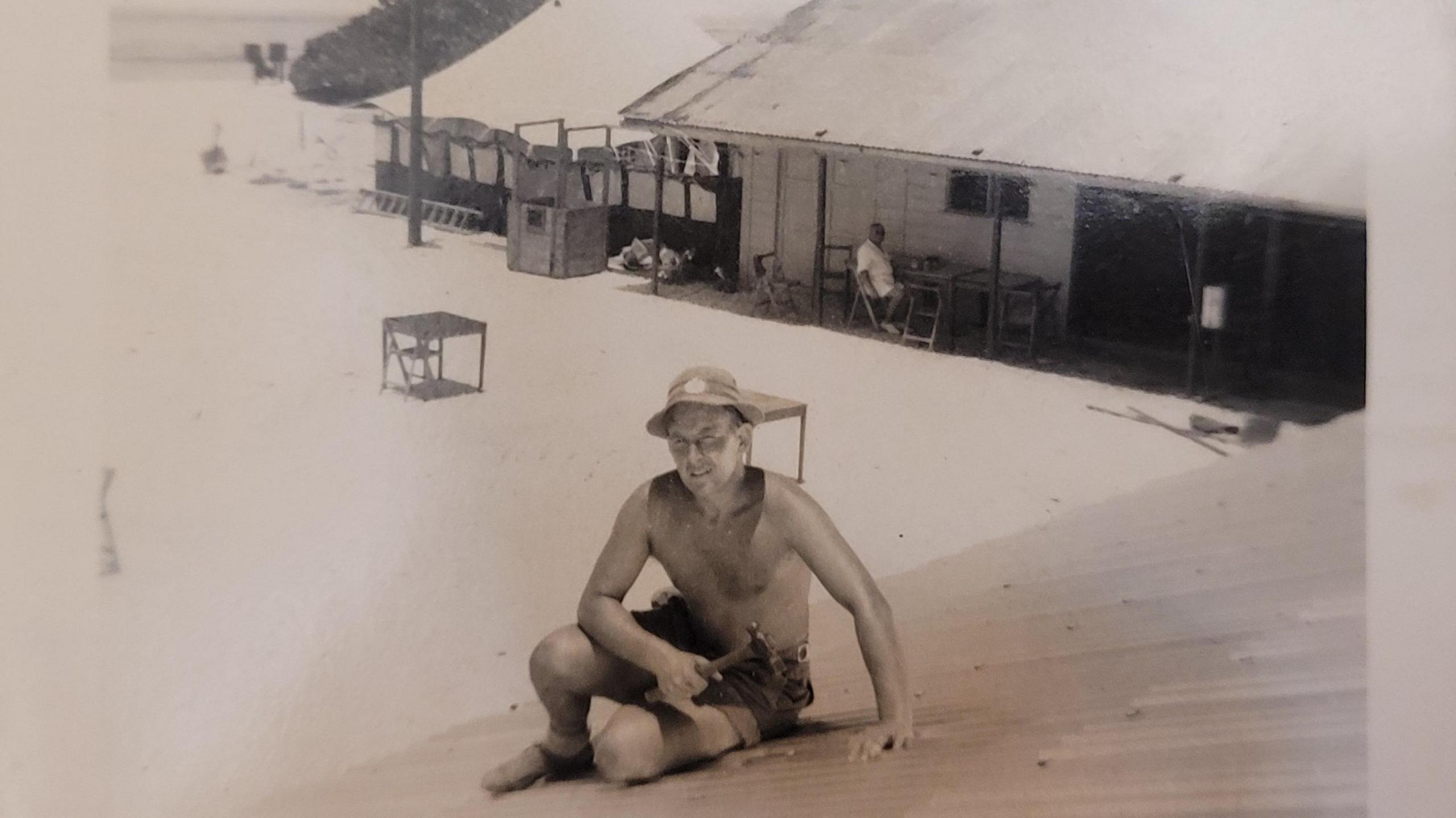 A sepia photo showing Robert Carman on a roof holding a hammer with a hut in the background