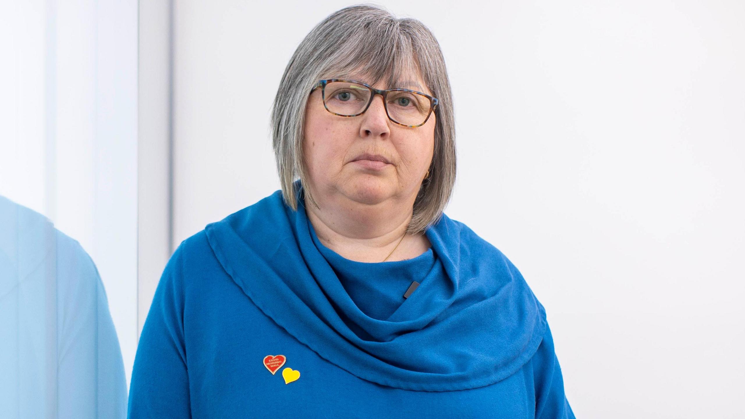 Brenda Doherty looking at the camera, she has grey hair and glasses and is wearing a blue dress with a heart pin. 