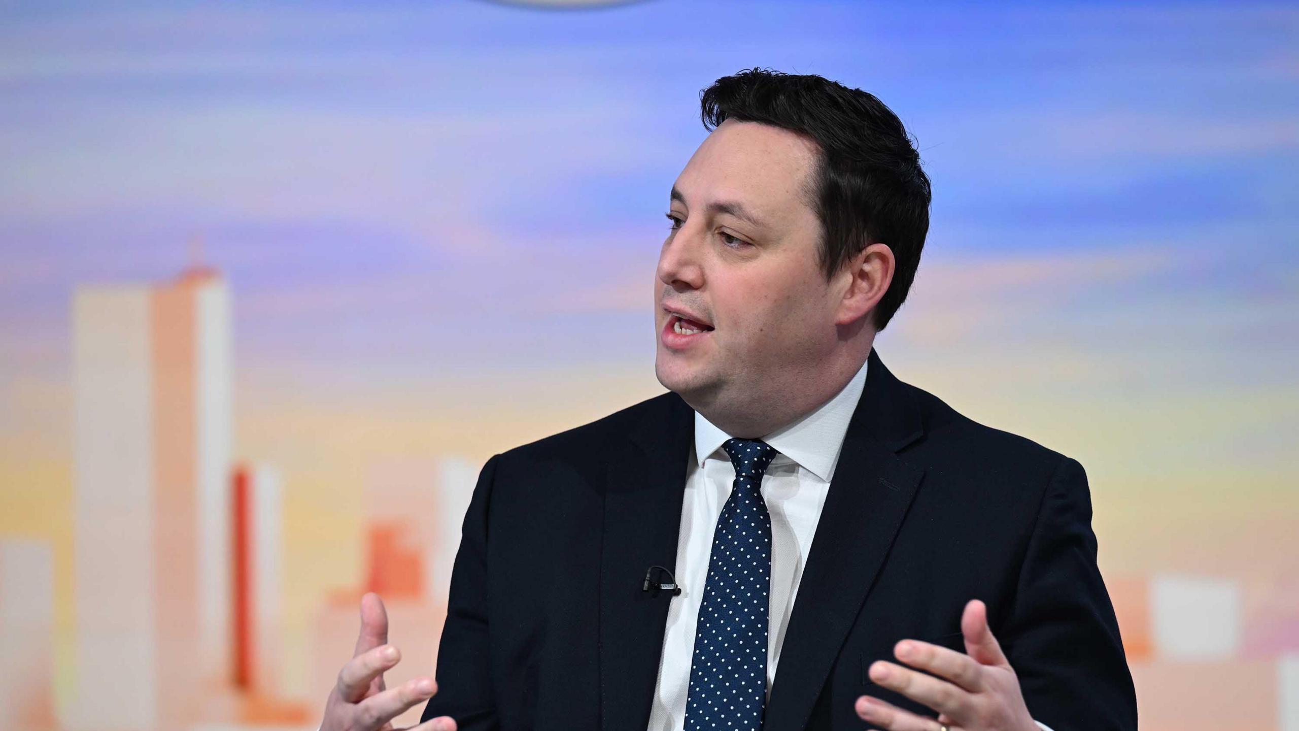 Ben Houchen. He has dark hair and is wearing a black suit and a blue tie with white spots. He is speaking on a BBC set in front of picture of a colourful skyline. He is gesturing with his hands while he speaks.