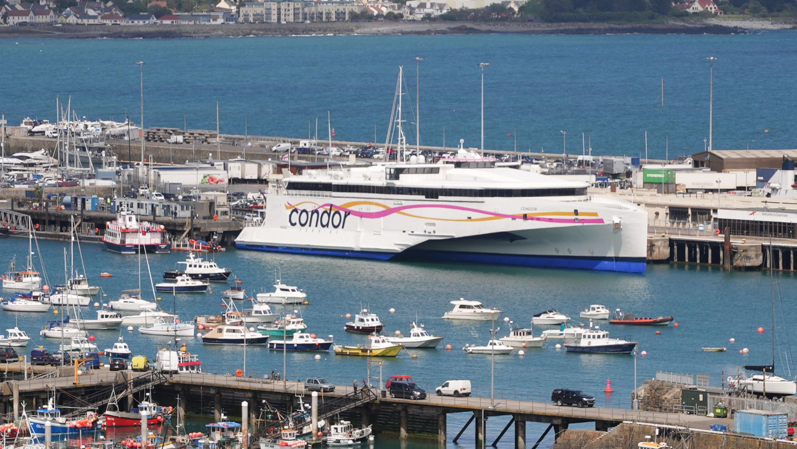 The Condor Liberation, a white catamaranm with the branding for Condor on the side, in St Peter Port Harbour surrounded by smaller boats. 