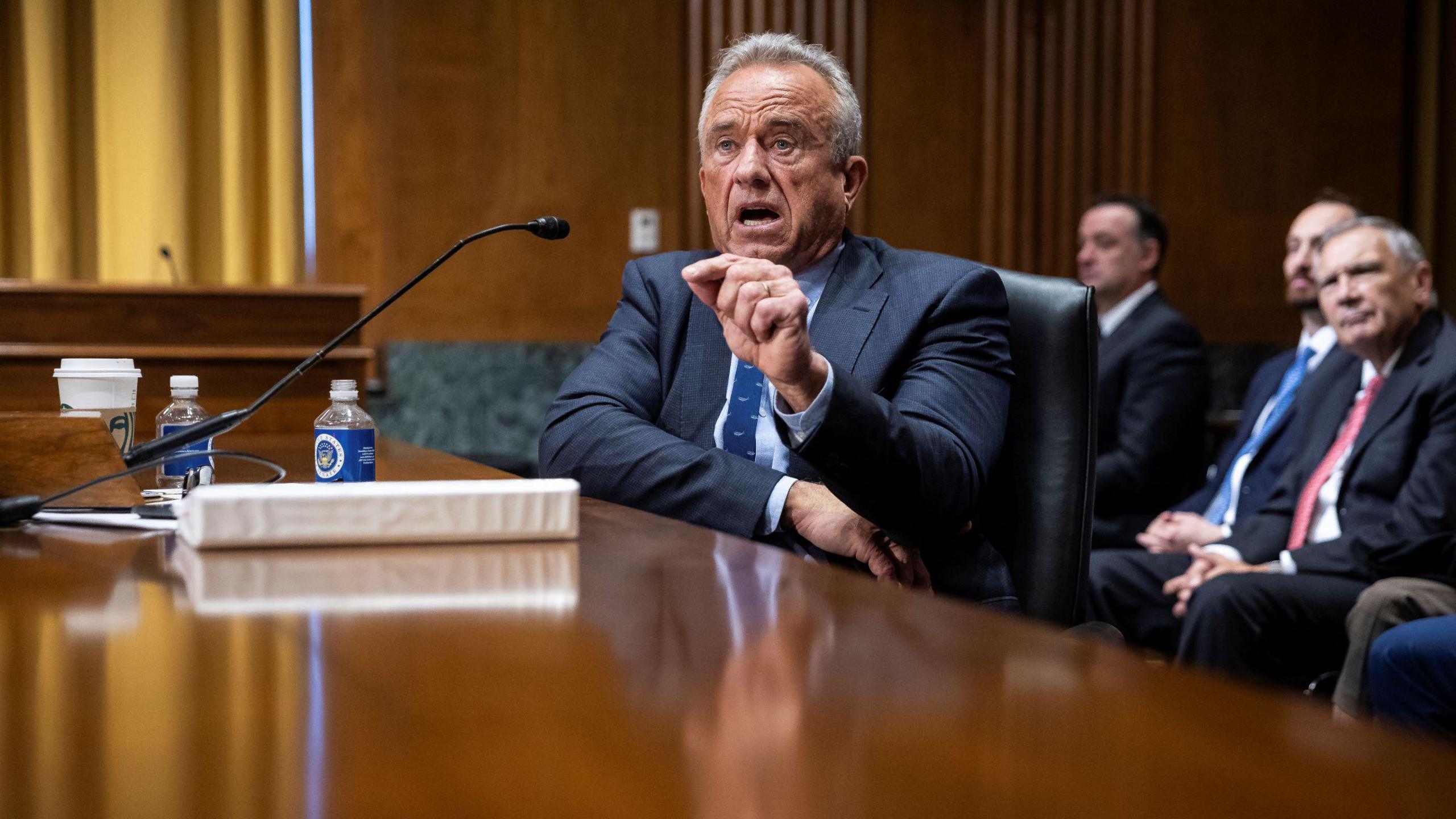 Robert F Kennedy, sits at a wide wooden table. He is wearing a smart, tailored, dark suit. On the table is white folder, some plastic bottles and a white disposable drinks cup. He is speaking animatedly and gesturing with his left hand. In the background, other men in similar dark suits listen. 
