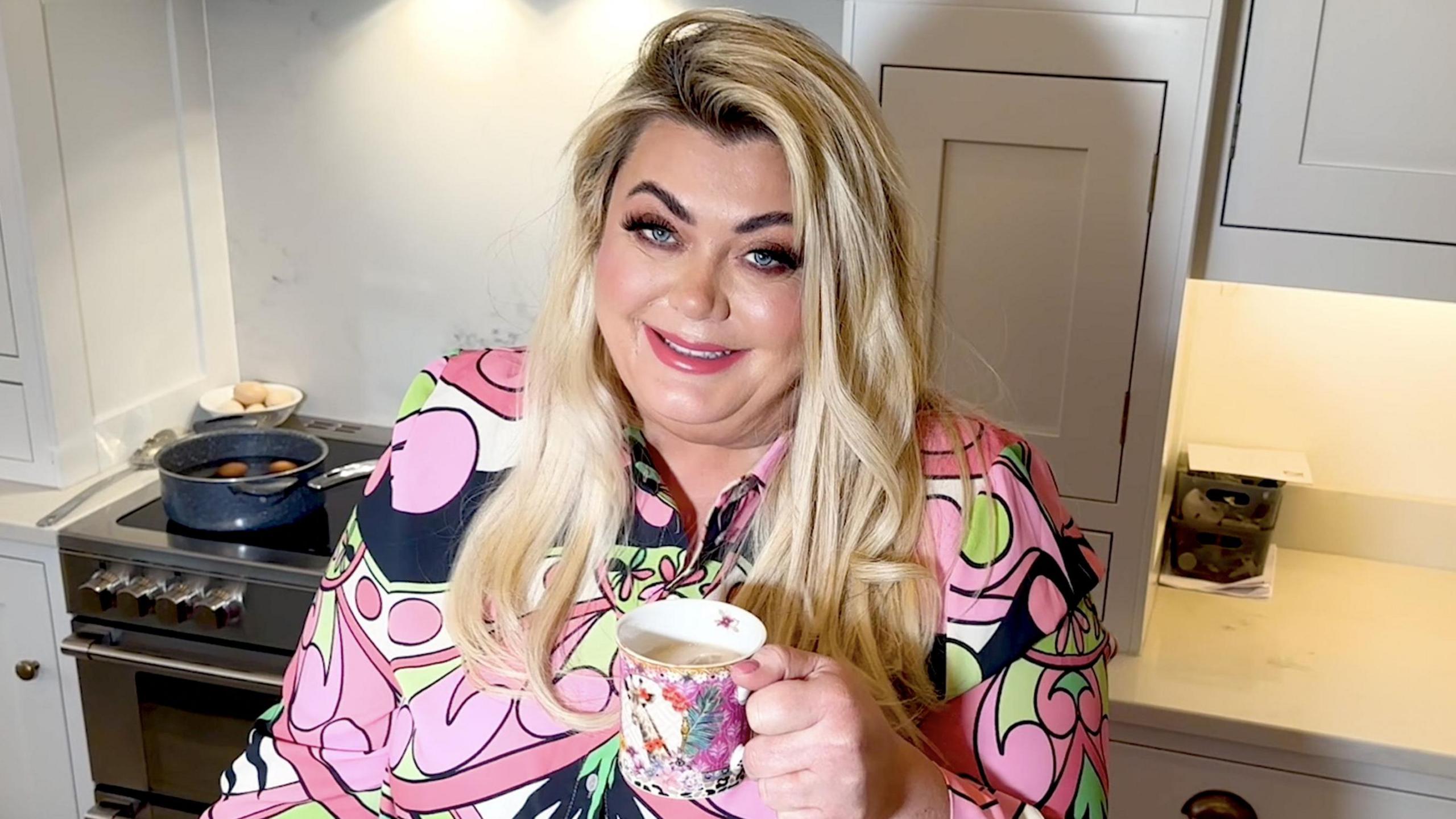 Gemma Collins stands in a kitchen holding a cup of tea and smiling