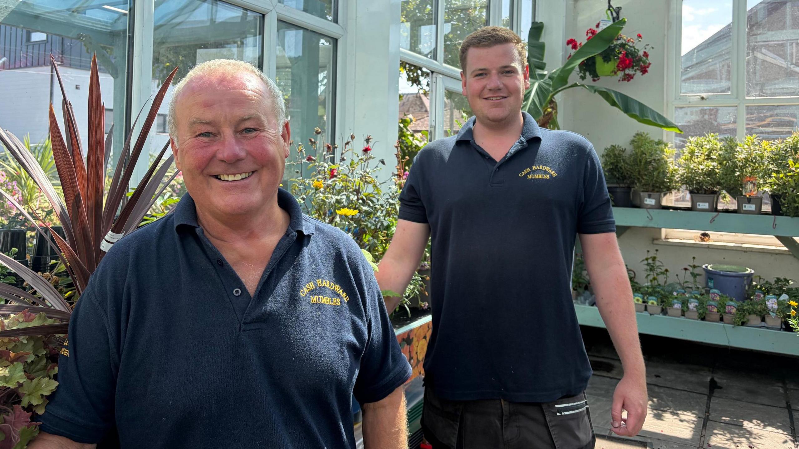 The two men with navy t-shirt standing in front of plants. Chris is older and stands in the front, with Alfie slightly behind him, both have short hair and are clean shaven.