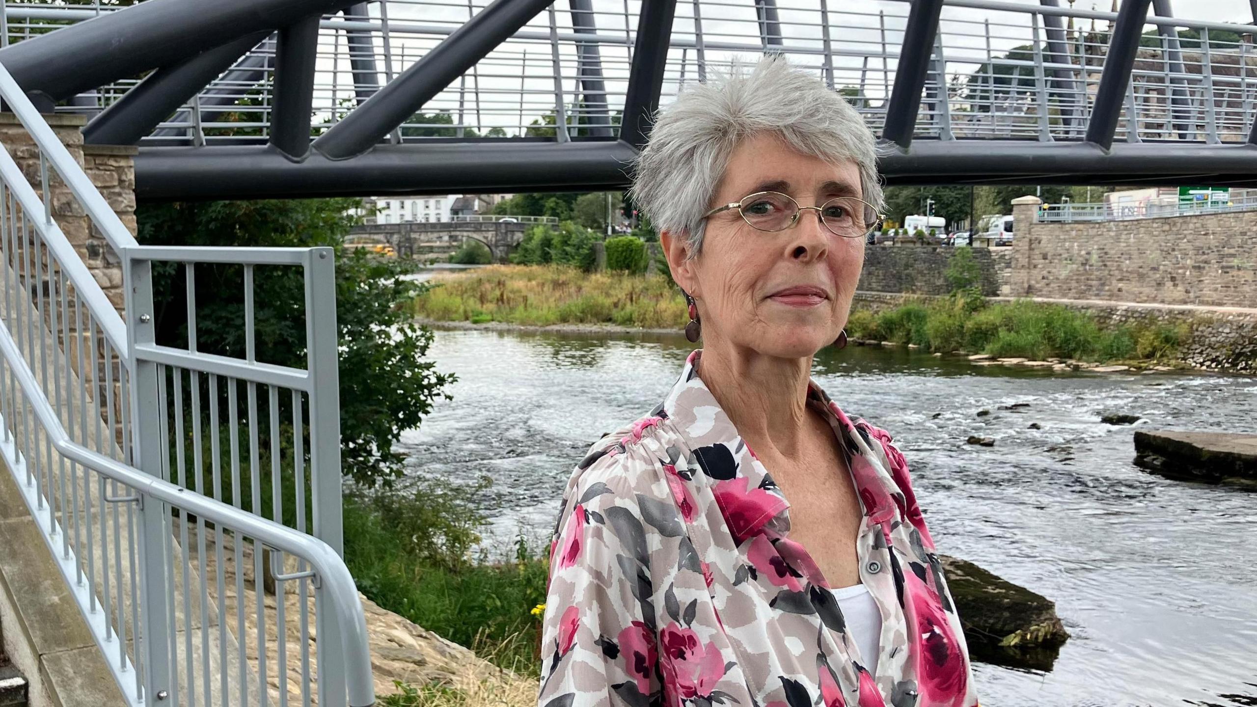 Lis Dales is standing in front of a foot bridge over the River Kent in Kendal. She is wearing glasses and a white and grey blouse with pictures of pink roses on it.