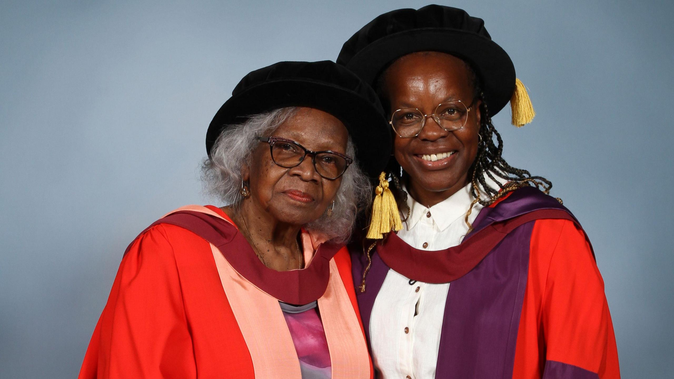 Mrs Dettering standing beside Dr Gournet and leaning her head towards her shoulder. They are both wearing red graduation robes and black graduation caps with yellow tassels. They are smiling and standing in front of a blue-grey photography background
