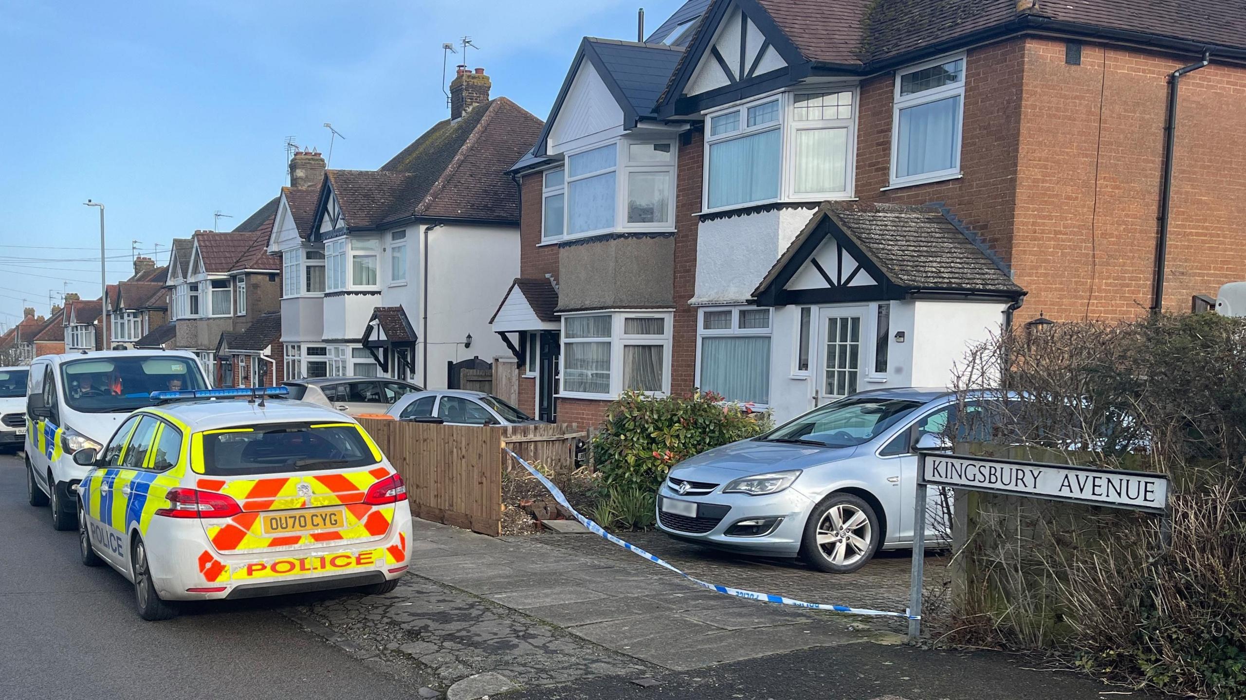 A marked police car - which is blue, yellow and orange - is parked outside a semi-detached red-brick house. There is police tape across the drive on which a car is parked. A street sign to the right of the house reads Kingsbury Avenue.
