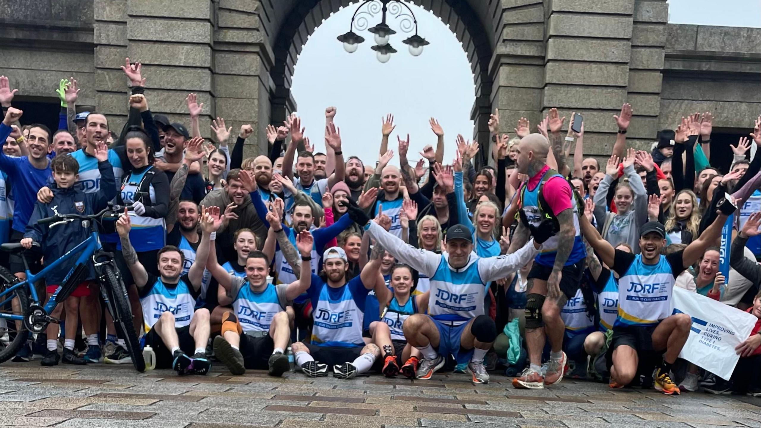 A group of about 60 runners sit and stand with their hands in the air in front of an archway. They are looking towards the camera and smiling.