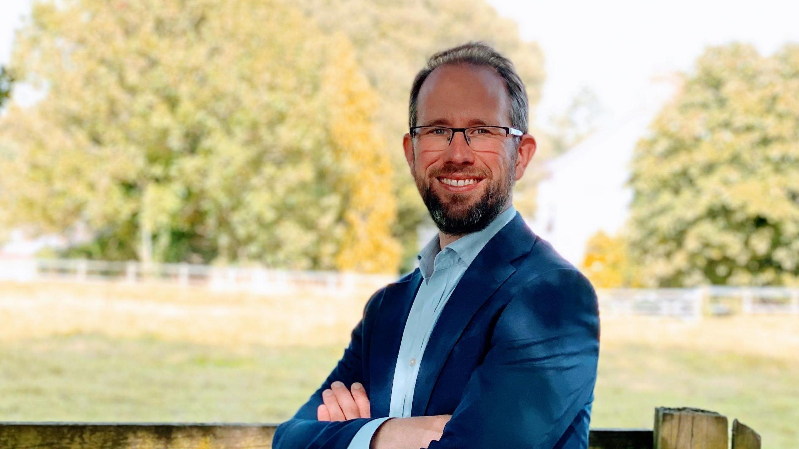 TV PCC Matthew Barber. He is wearing a blue blazer and light blue shirt, and has a brown beard and glasses. He is stood in front of a grass field.