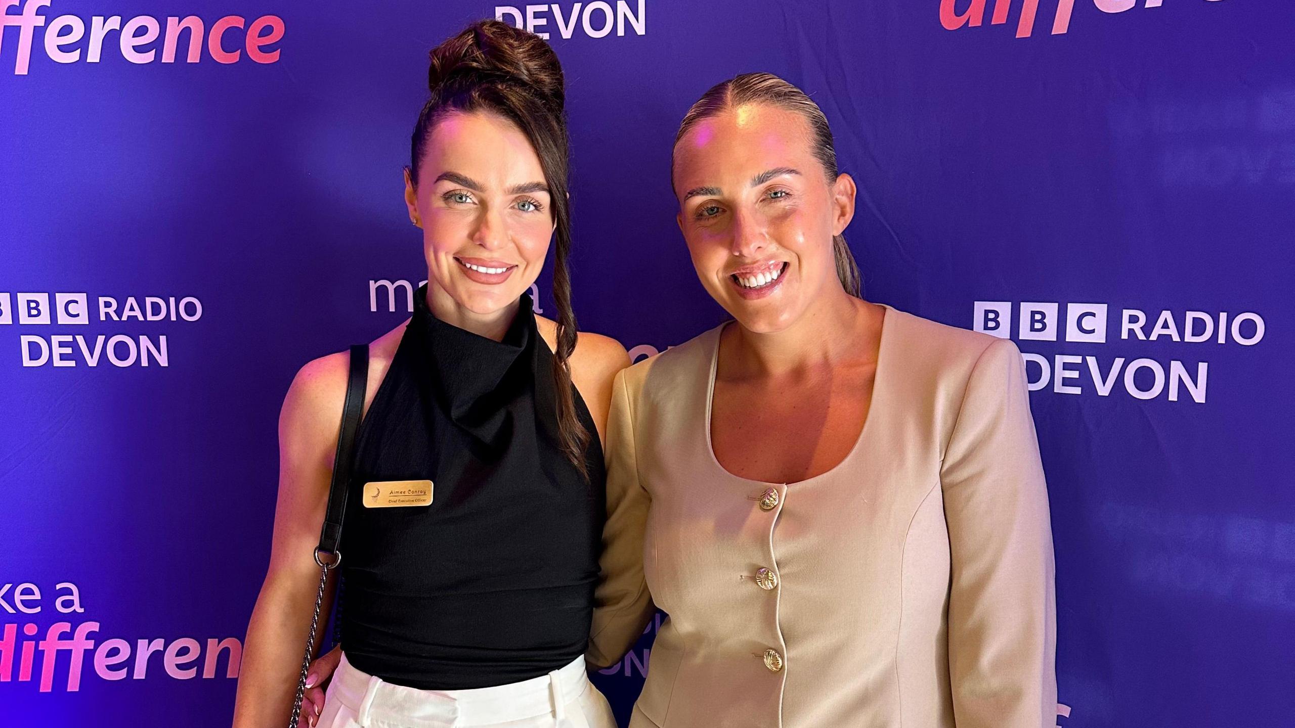 On the left is Aimee Conroy wearing a high-neck black top with white trousers. She has brown hair which is tied up. On the right is her guest who is wearing a beige dress with buttons down the front.