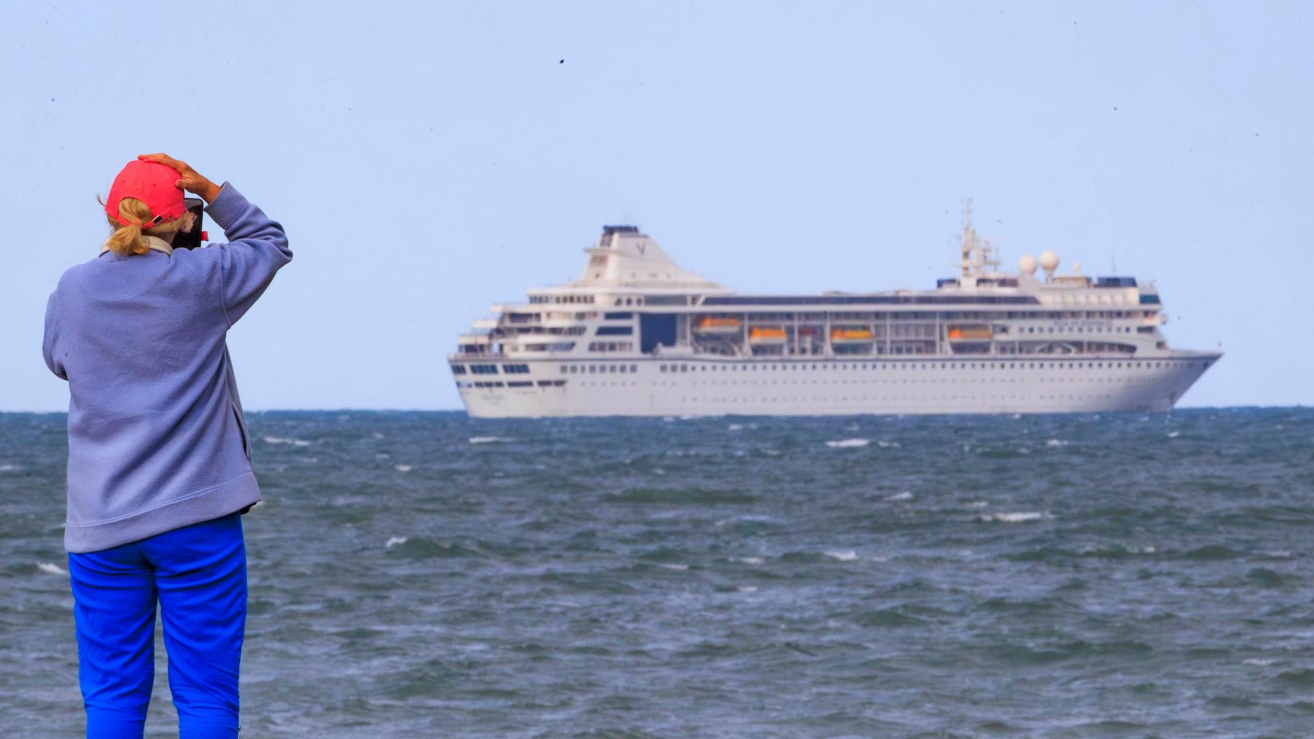 A person wearing a red cap, light blue fleece and bright blue trousers is taking a photo of the cruise ship with a phone. The ship can be seen in the distance with the yellow lifeboats on display on the side of the white ship.