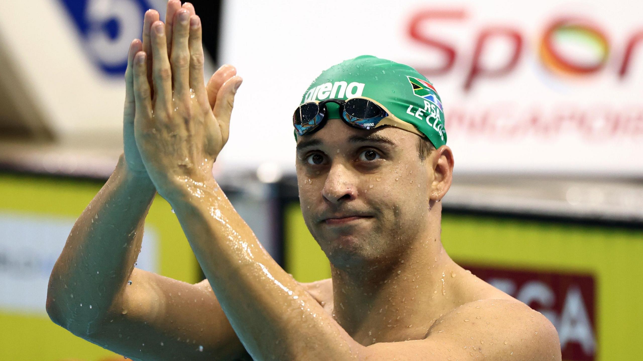 Chad le Clos holds his hands together in a prayer-like gesture above his head in a swimming pool as he wears a green South Africa swimming cap and goggles around his forehead. Sponsors' logos and a set of starting blocks can be seen out of focus in the background