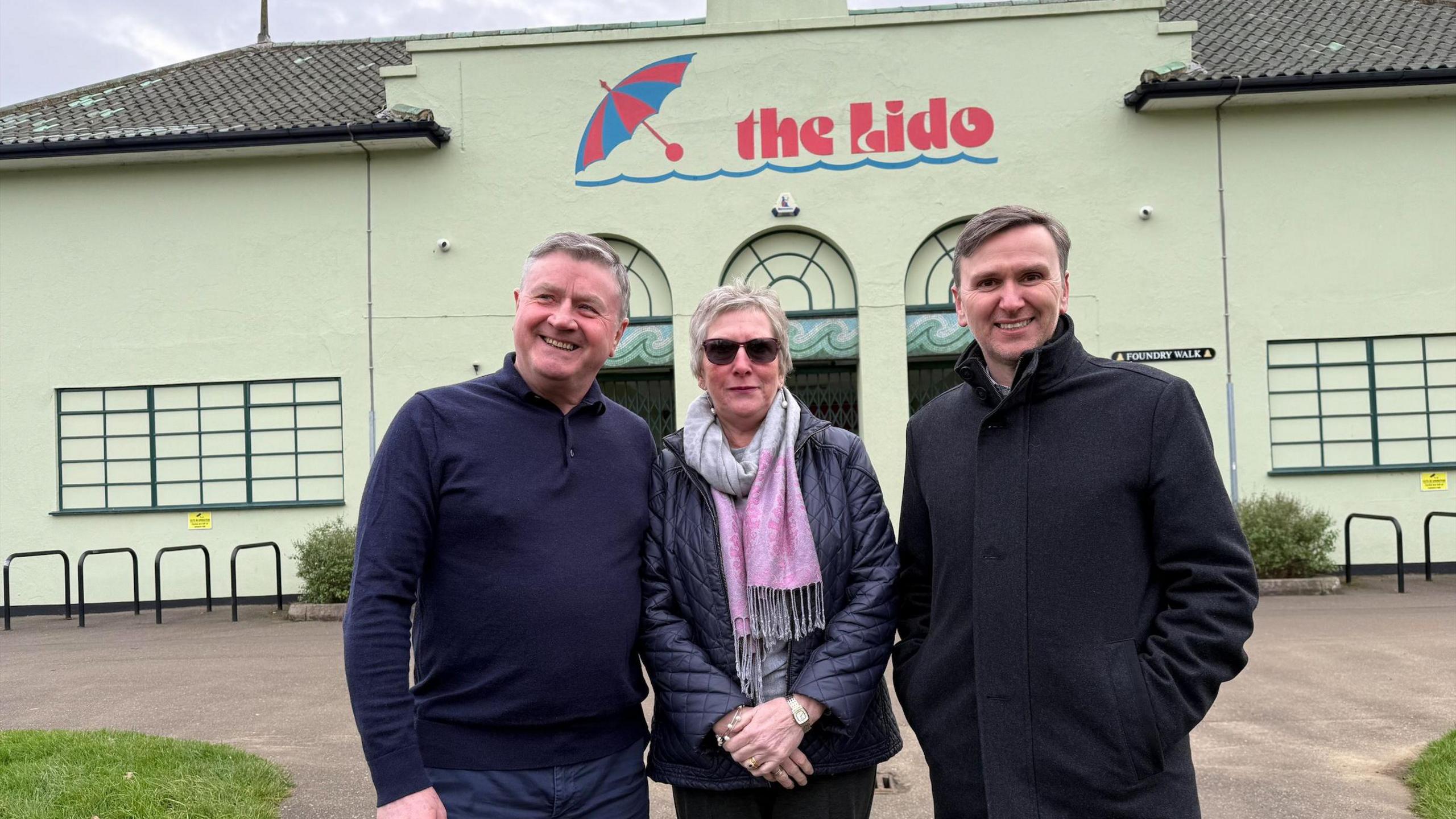 A man wearing a navy knitted top standing next to a woman wearing a blue padded jacket and a pink and grey scarf. On the right is a man wearing a black coat with his arms in his pockets. Behind them is the outside wall of the lido, with the words "The Lido" written on it in red