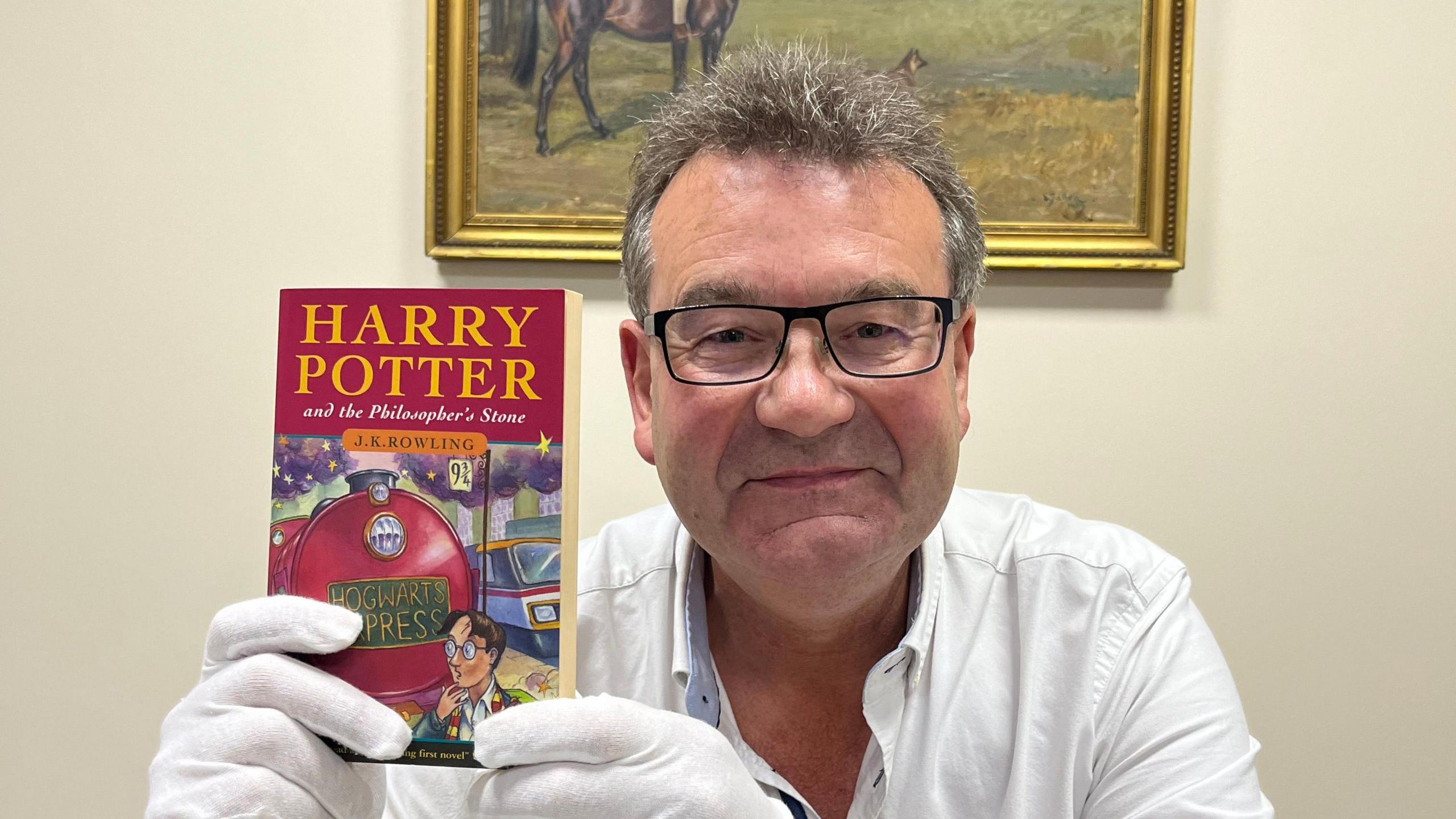 Richard Winterton, who has short dark hair and wears black-rimmed glasses, holding the Harry Potter book. He is wearing white gloves and a white shirt. 