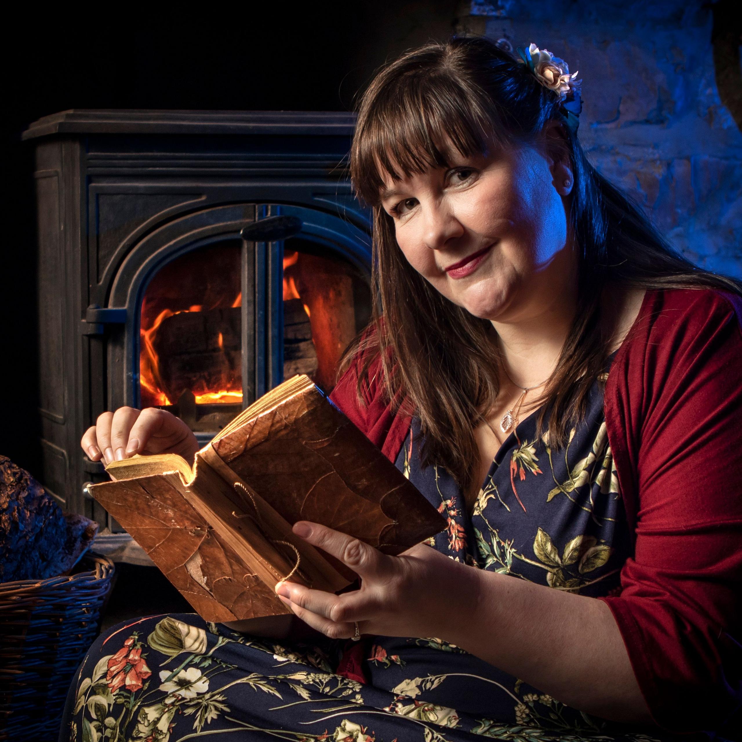 A woman with long dark hair reads from an old book in front of a roaring fire