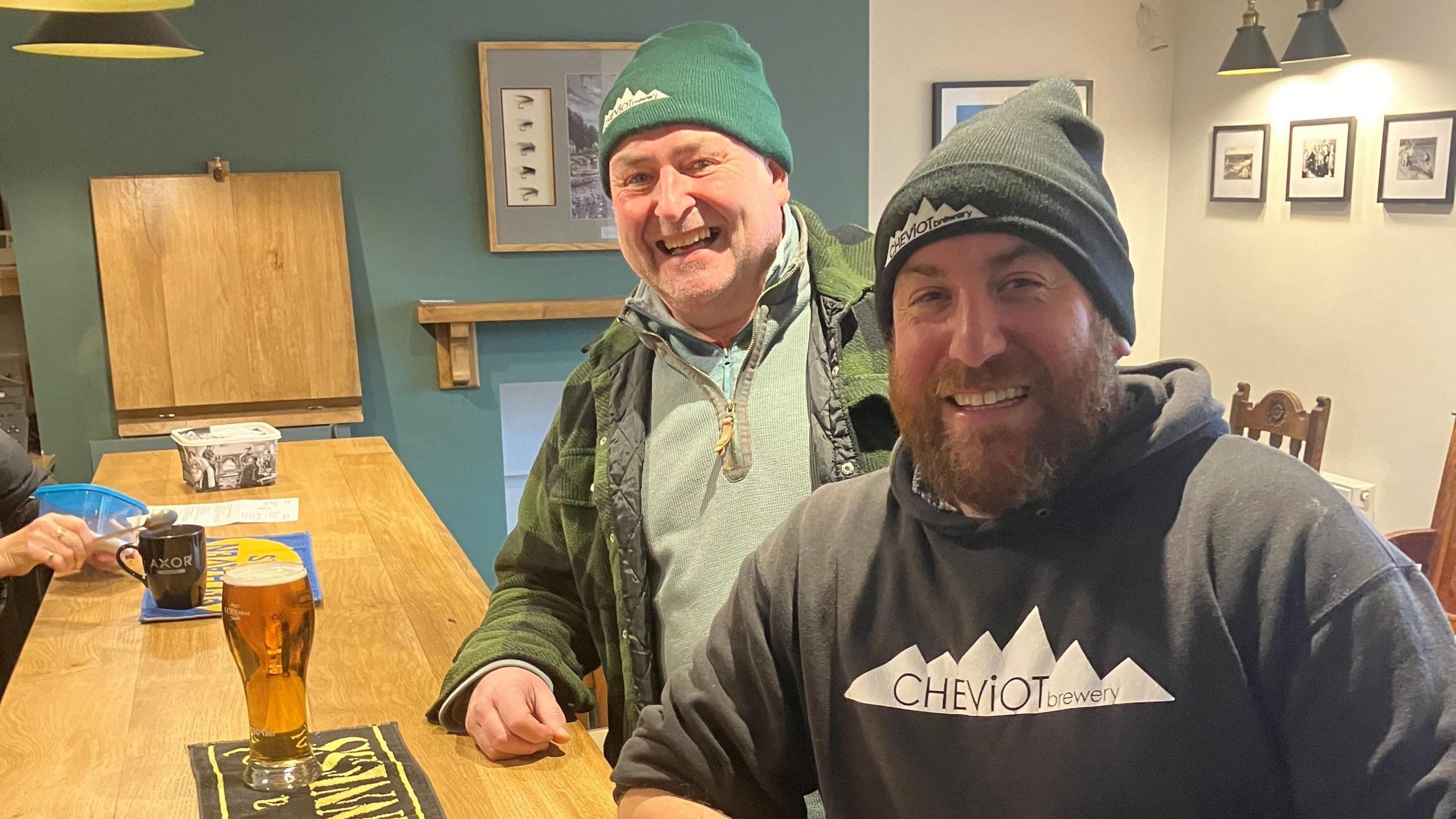 Two drinkers in woolly hats at the bar smiling broadly and ordering pints.