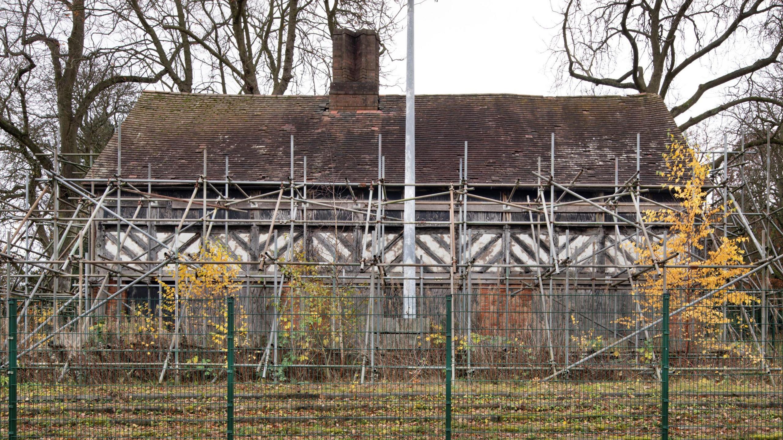 An old black and white pub building surrounded by scaffolding and behind a green fence and a number of trees and plants.