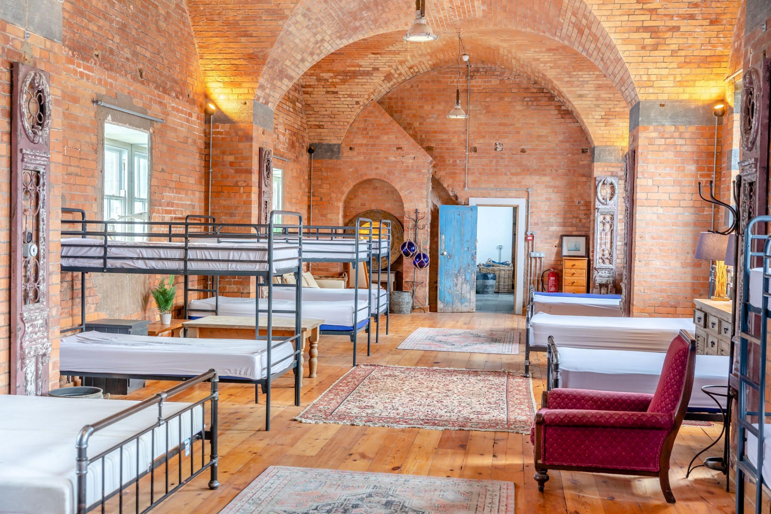 A room with exposed red bricks and three sets of bunk beds in a row, all with white sheets on. In a second row there are three single beds, and a red armchair closest to the camera. 