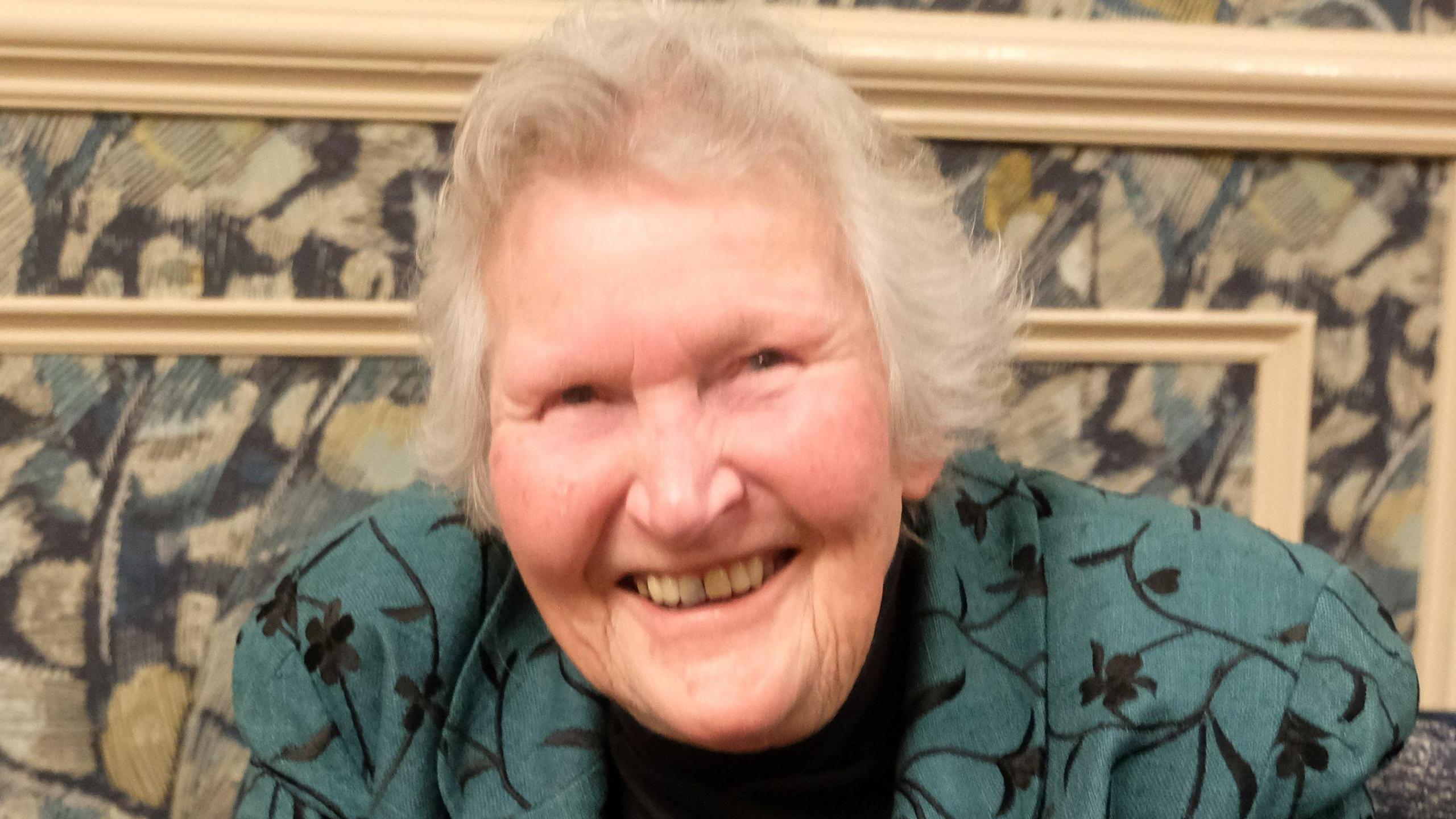 Gill Green, smiling to the camera, sitting down, wearing a black top, cat broach and a green and black jacket. She has short grey hair.