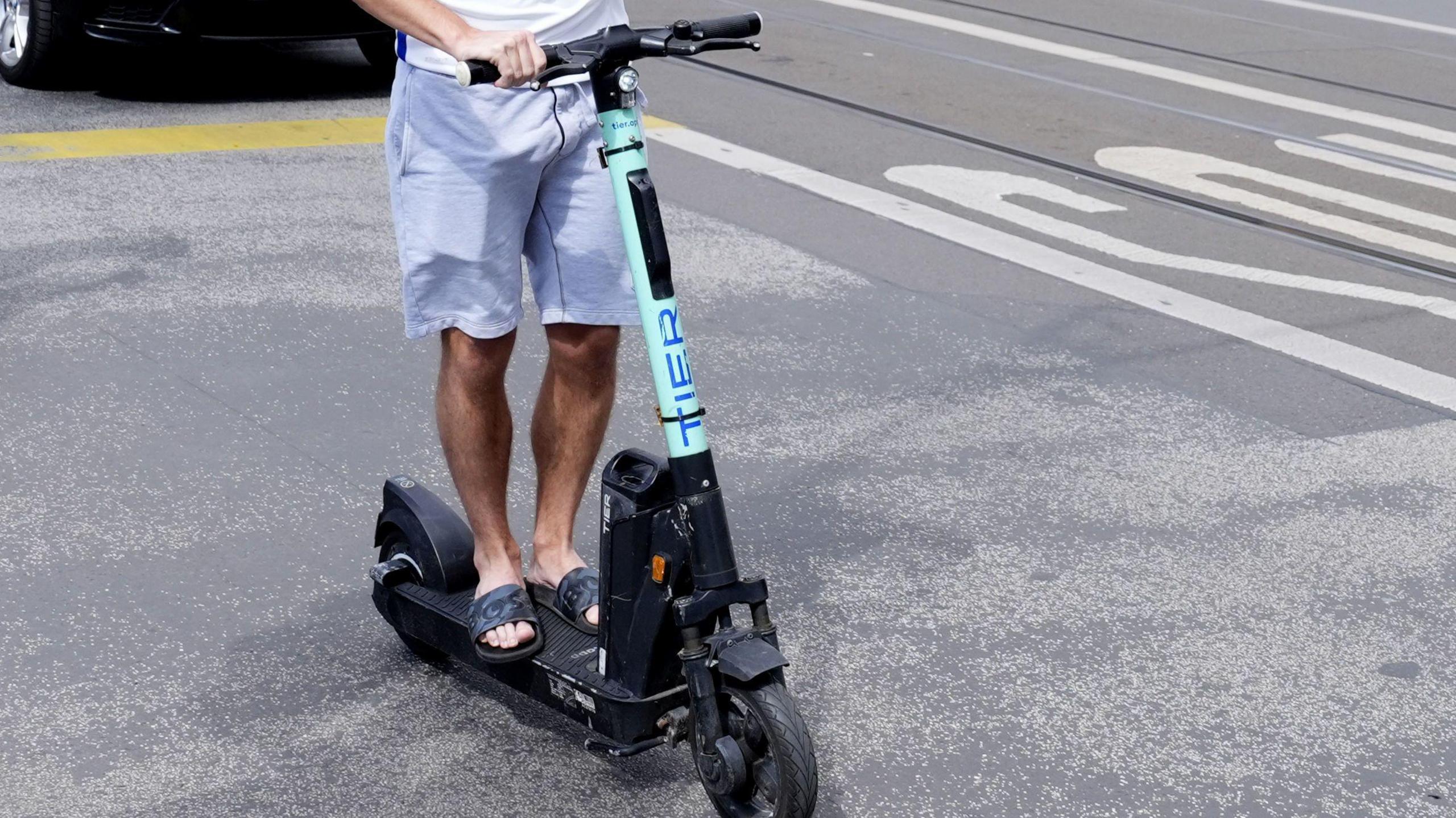 What appears to be a man on an e-scooter wearing flip flops and a pair of shorts. The scooter is blue and black. 