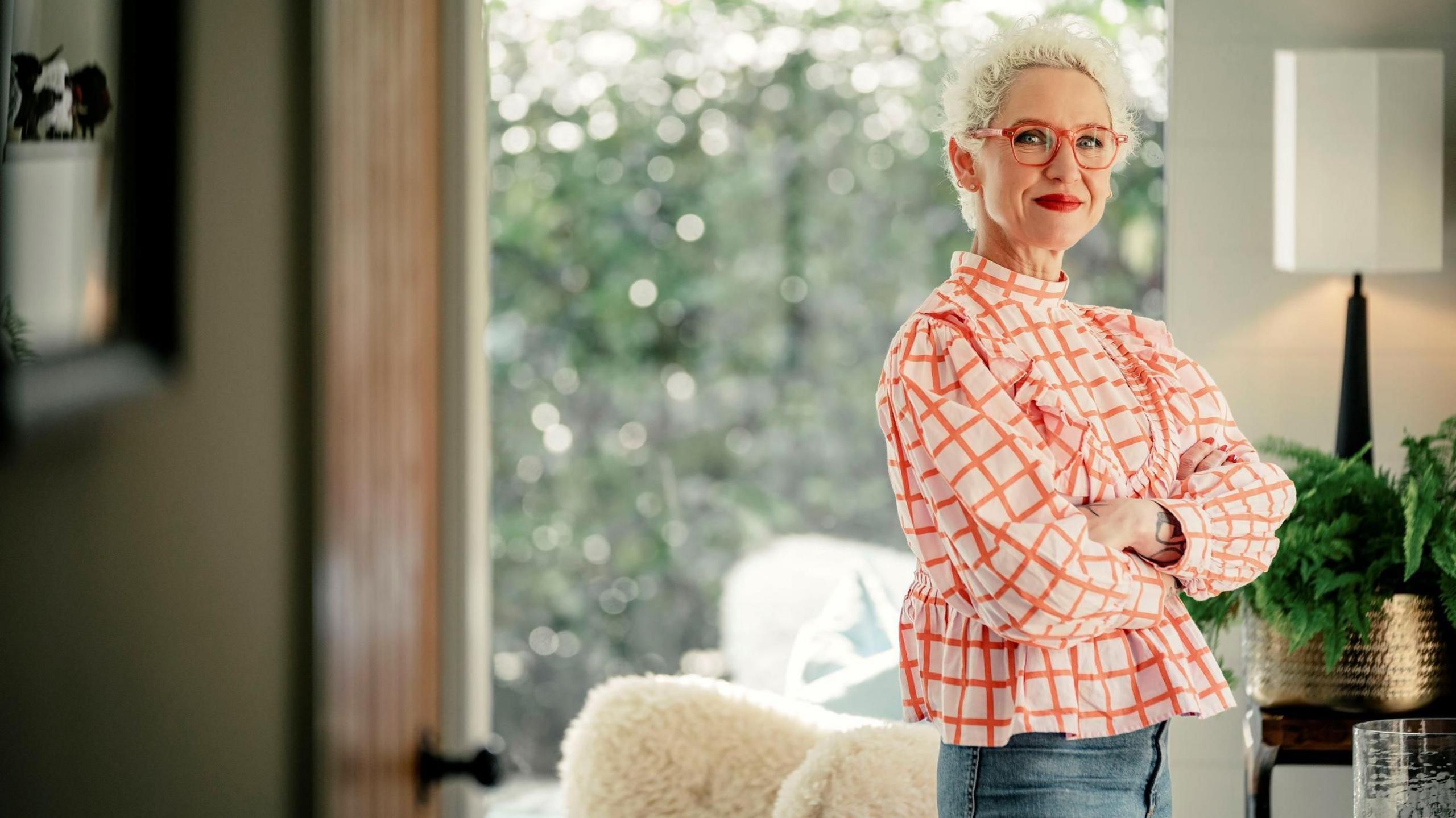 SHOTY judge Anna Campbell-Jones stands side-on, her arms folded, smiling at the camera in a bright airy living room. There is a chair with a fluffy throw beside her, a table with a big lamp and a huge gold plant pot and a floor-to-ceiling window at the back shows us a leafy garden.