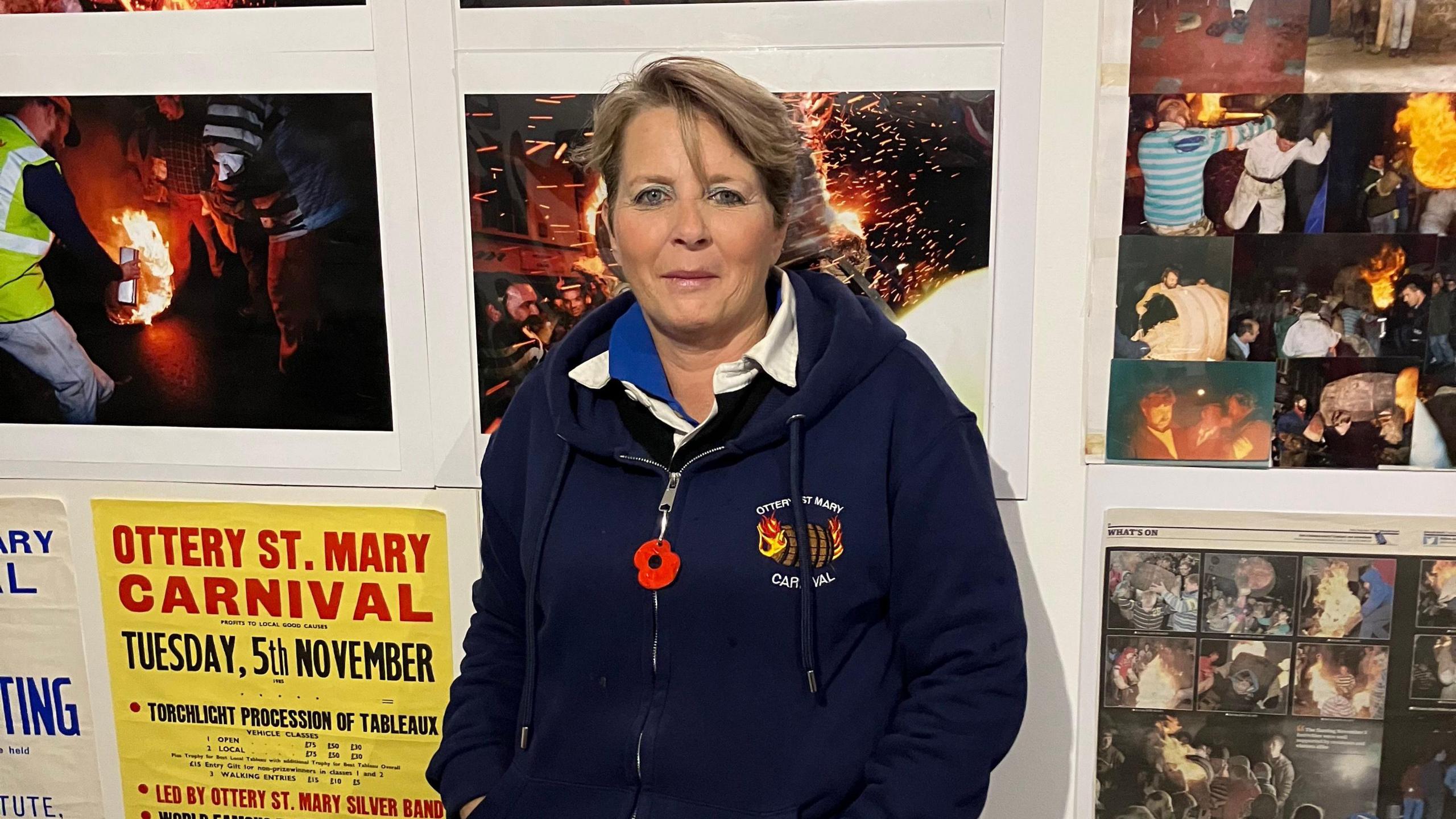 Carol Adshead-Wotton standing against a backdrop of photos from previous years of the event. She has short hair and is wearing a navy blue hoodie.
