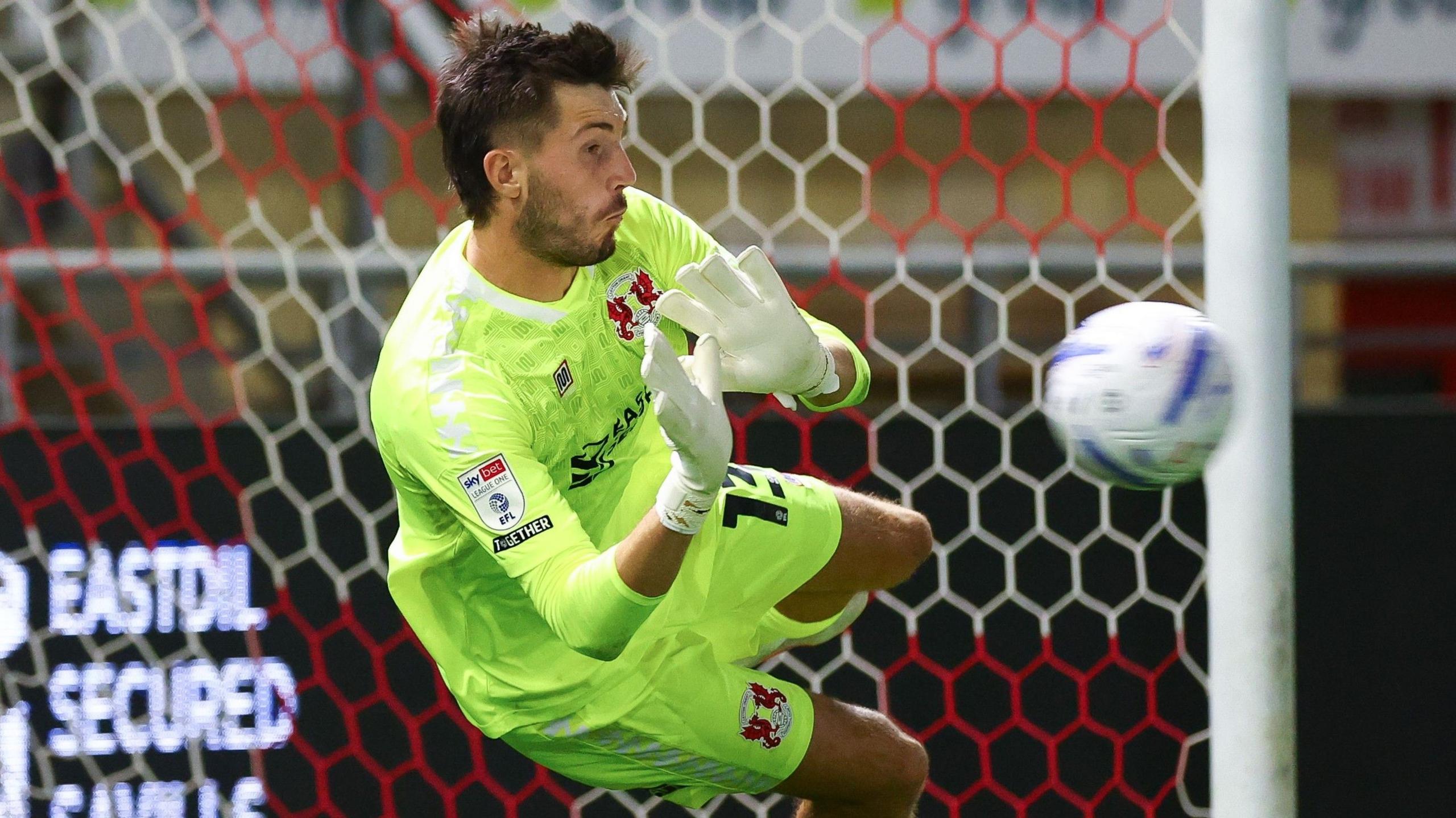 Leyton Orient goalkeeper Noah Phillips, wearing a light green shirt and white gloves, dives to his right to push away a penalty kick against Walsall