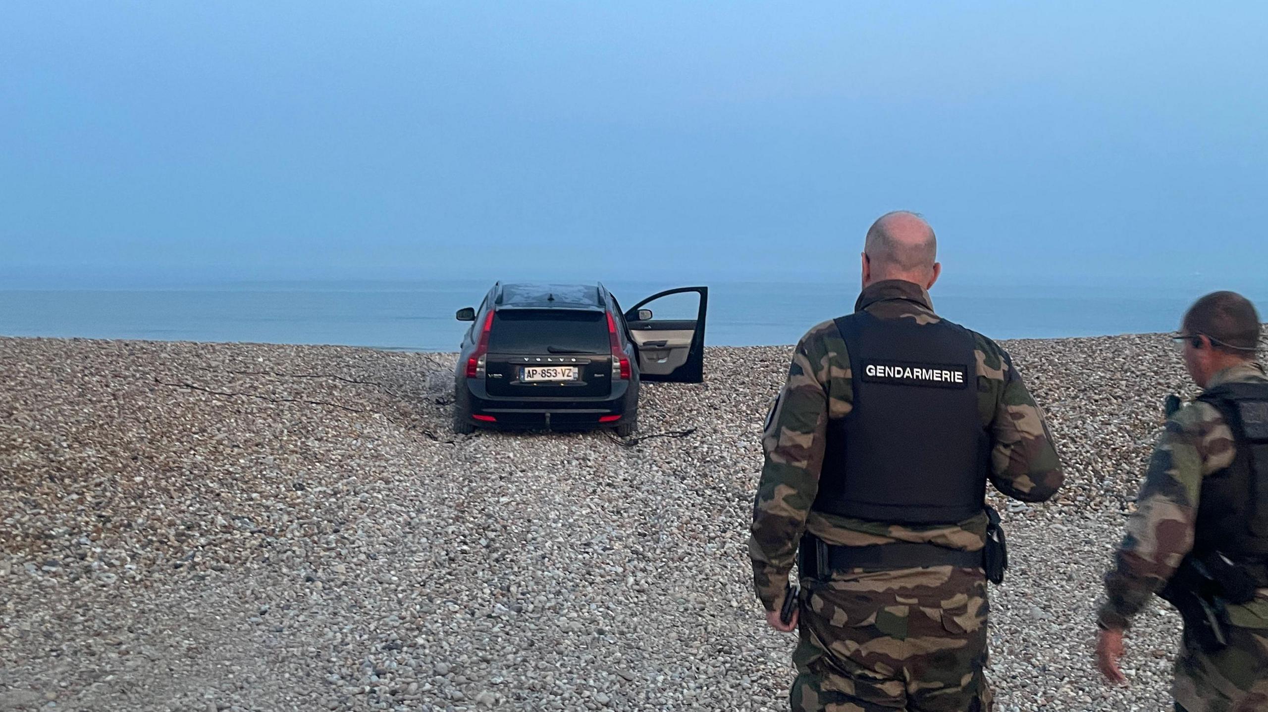Two police officers walk towards an abandoned black car sitting on a shingled beach, one door still open