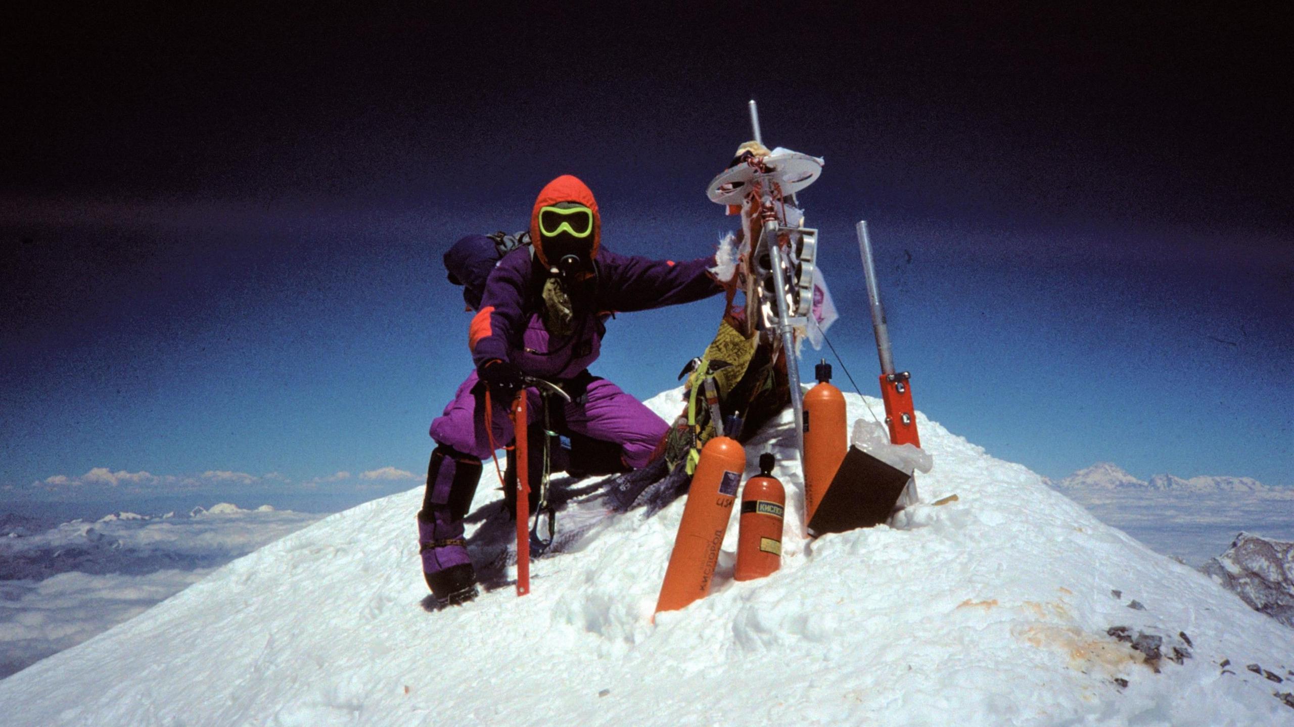 Caradoc dressed head to toe in purple mountain kit at the top of a snowy mountain, with clouds in the background and bright orange oxygen tanks in the snow in front of him.